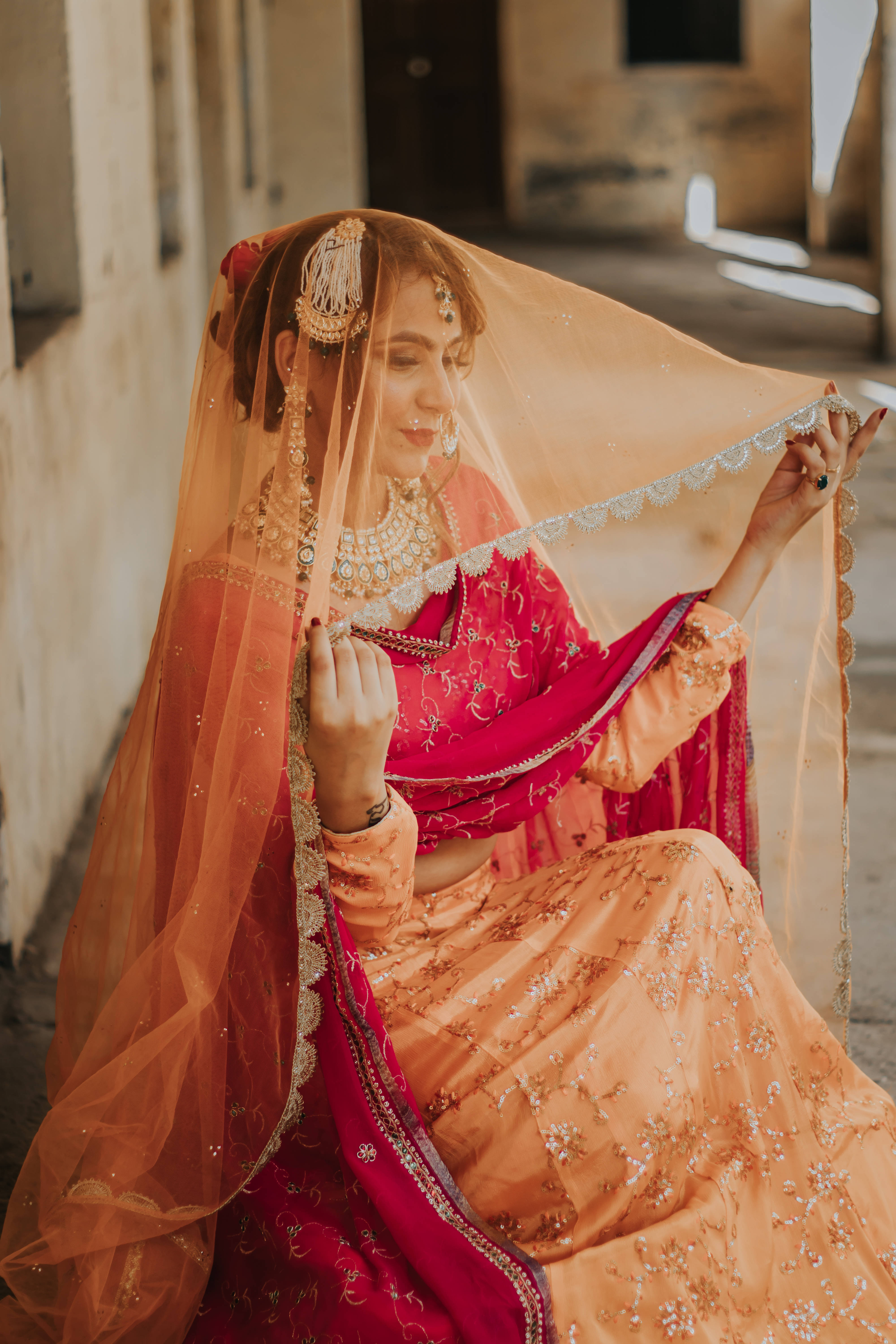 Indian 桌布 壁紙 — 一位身穿鮮豔紅桃色刺繡長裙、披著薄紗的女性，坐在陽光灑落的小巷中，溫柔微笑。