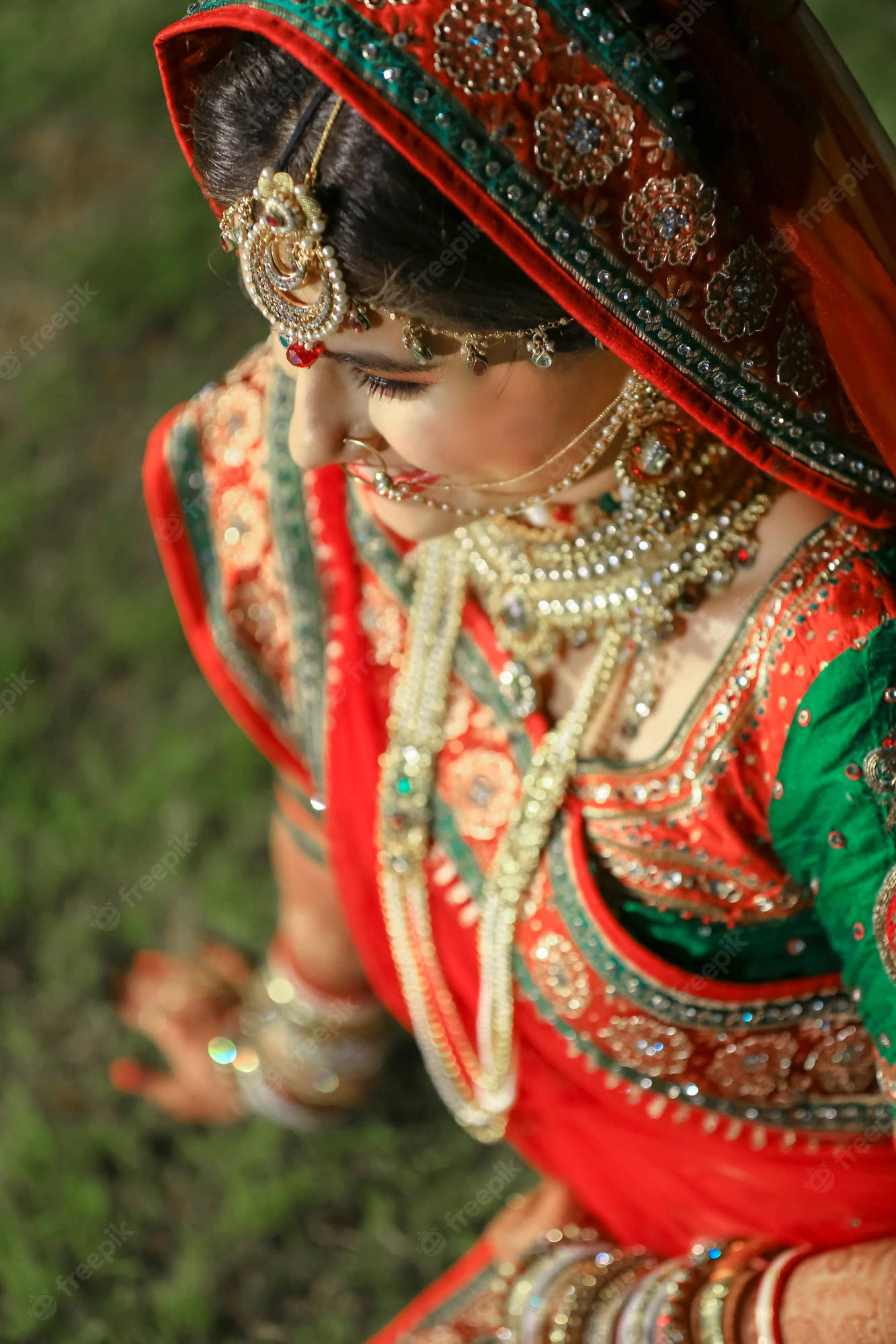 Bridal 桌布 壁紙 — 一位身穿鮮豔紅綠色傳統服飾、飾有精緻金飾和珠寶頭飾的女子，在柔和的綠色草地背景下低頭凝視。