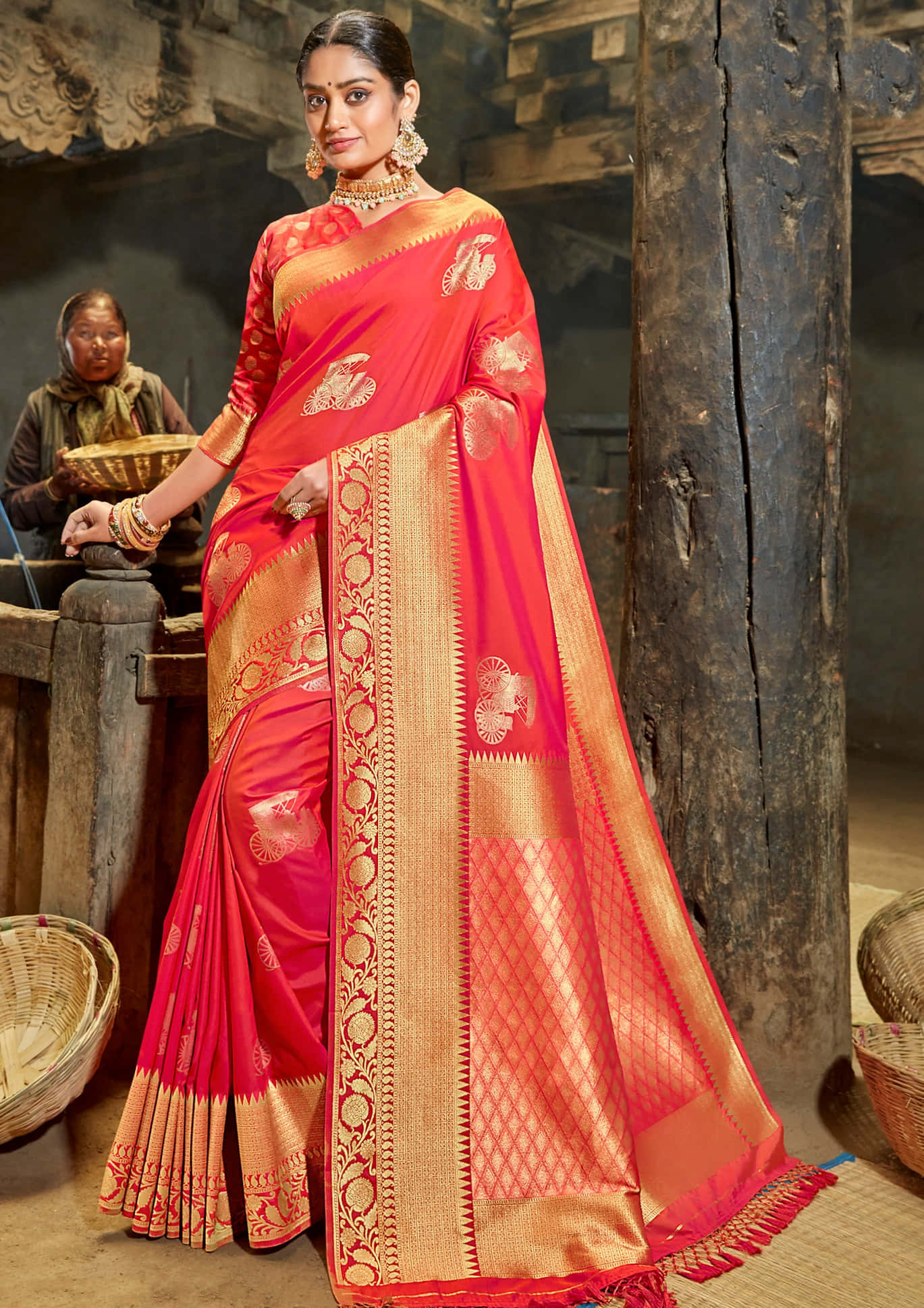 Saree 桌布 壁紙 — 一位身穿鮮豔紅色和金色刺繡紗麗的女性，在質樸的木質室內優雅地站立，背景有傳統編織籃子和一位坐著的女性。