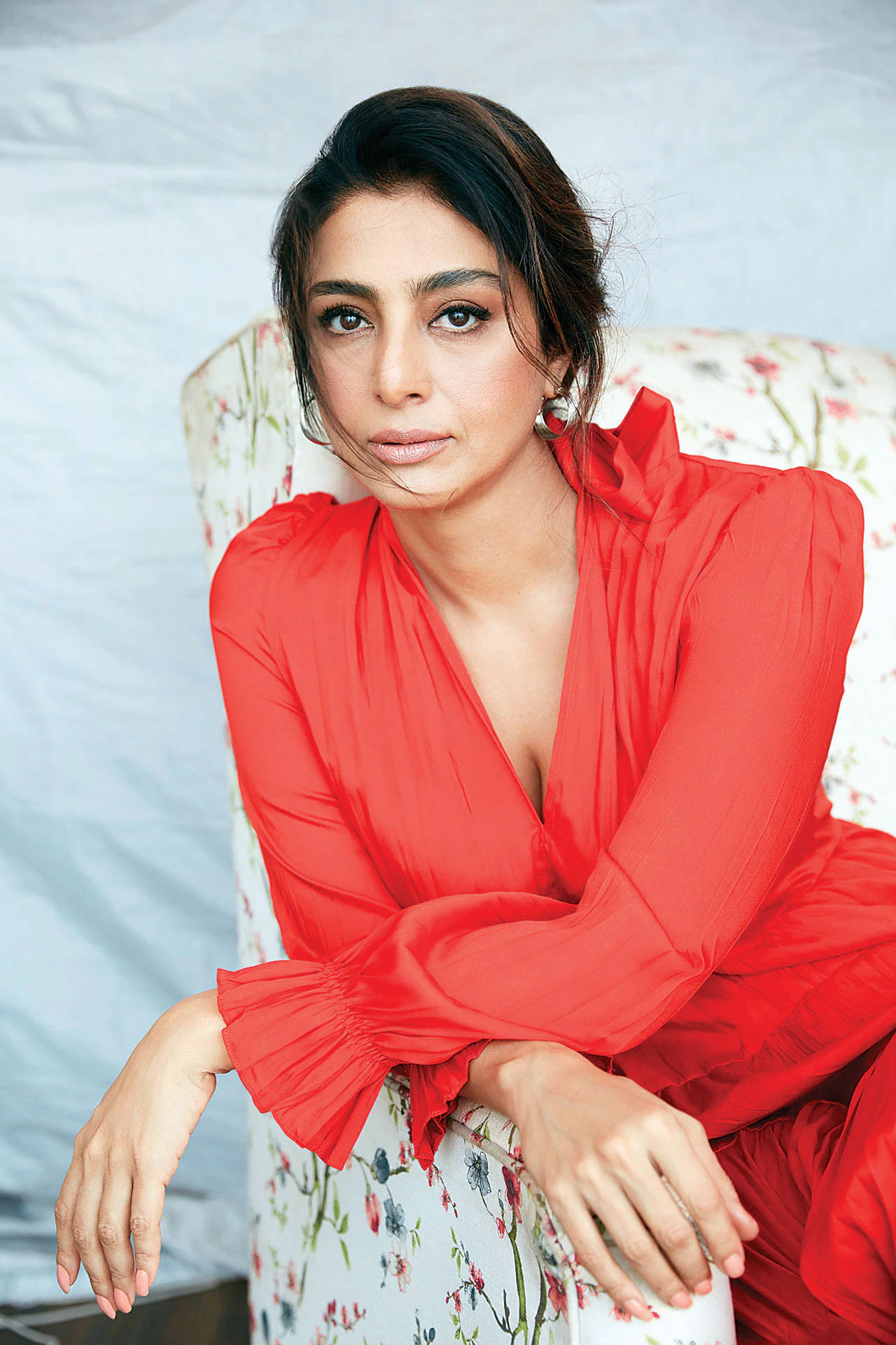 Indian Women 桌布 壁紙 — 一位身穿鮮豔紅裙的女性，優雅地坐在花卉圖案的椅子上，背景是柔和的灰色。