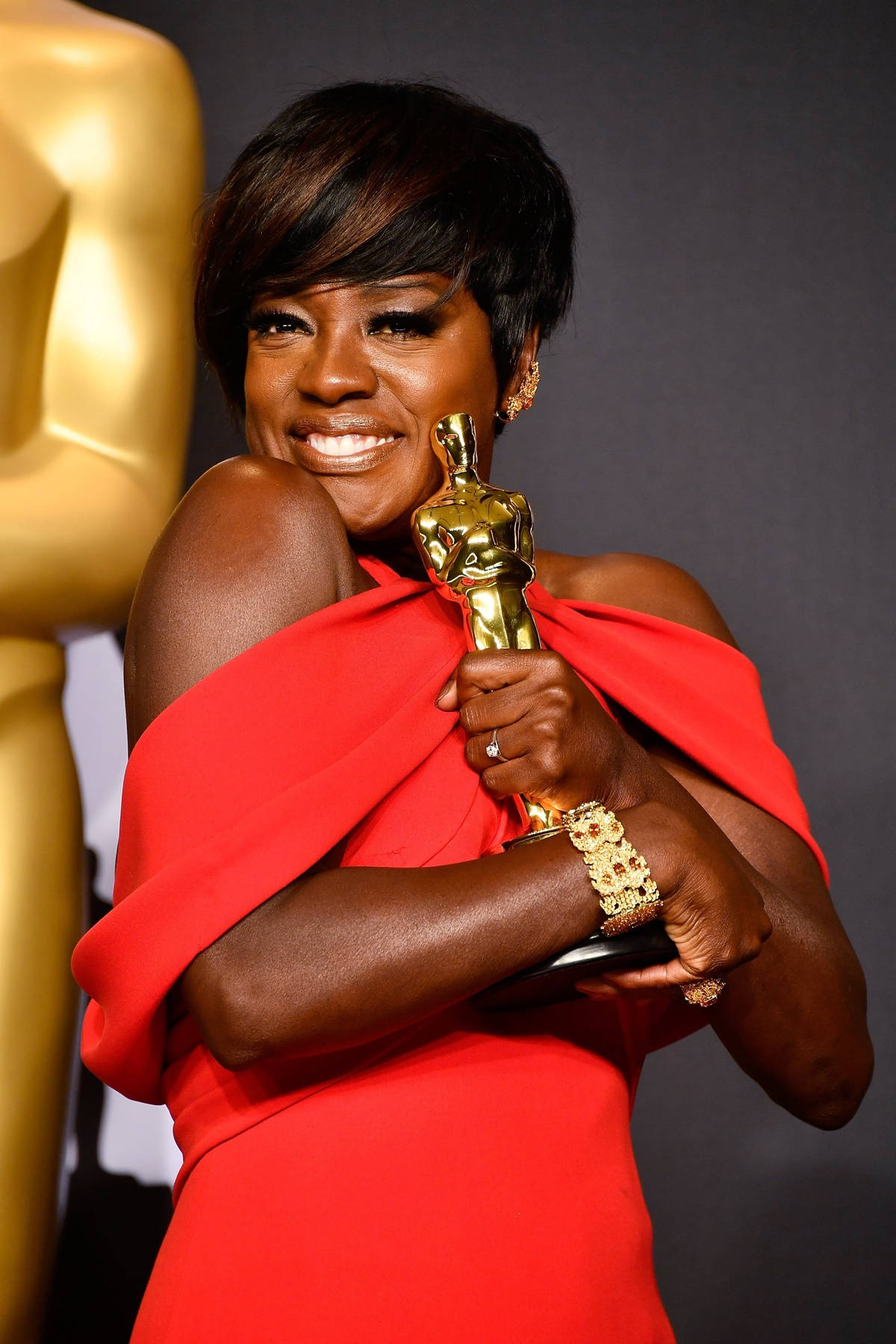 oscars 桌布 壁紙 — 一位身穿鮮豔紅裙的快樂女性，在黑暗背景下手持金色奧斯卡獎座，笑容燦爛。