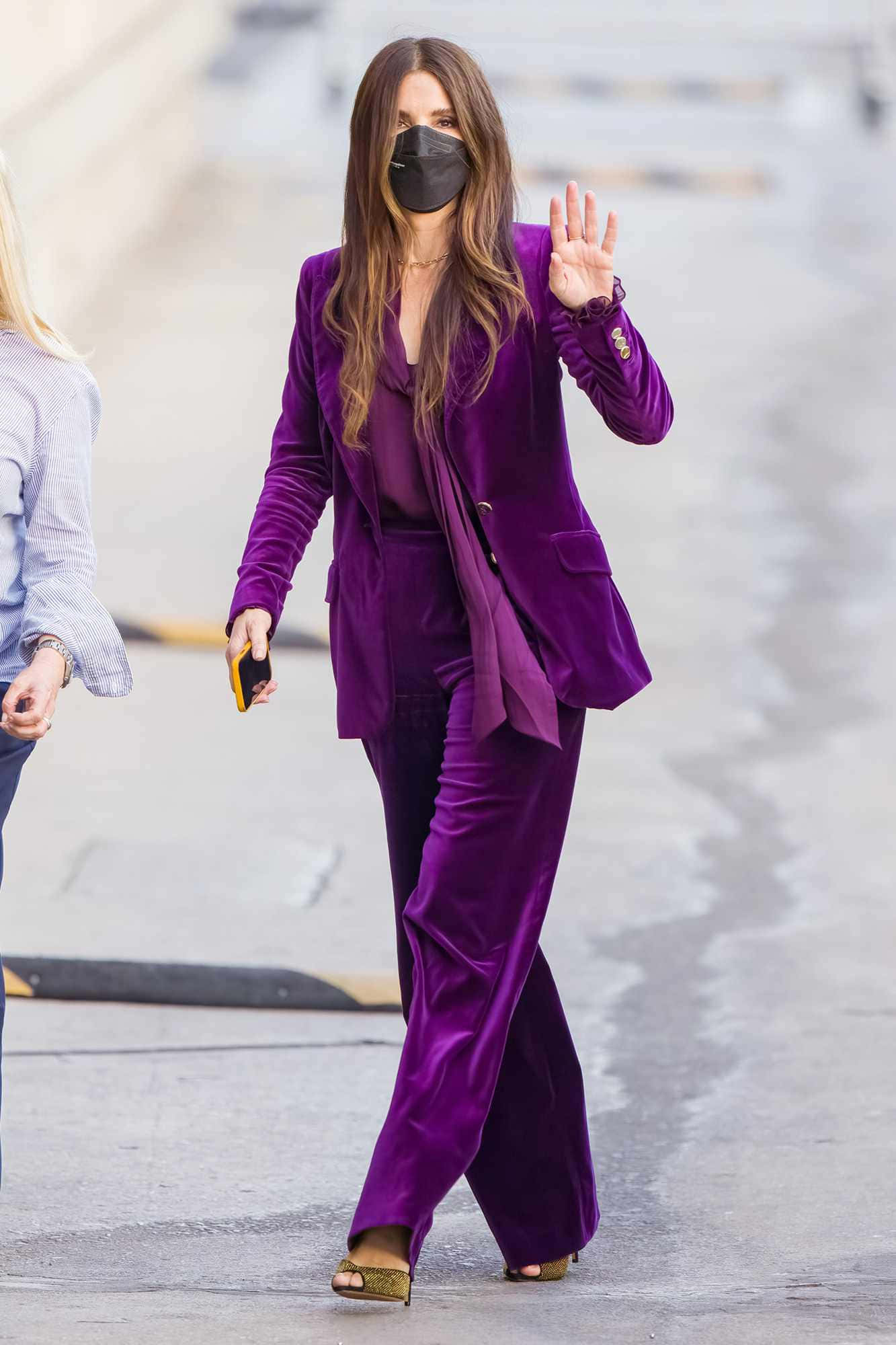 Sandra Bullock 2021 桌布 壁紙 — 一位身穿鮮豔紫色天鵝絨套裝的女性在戶外潮濕的人行道上行走時揮手。