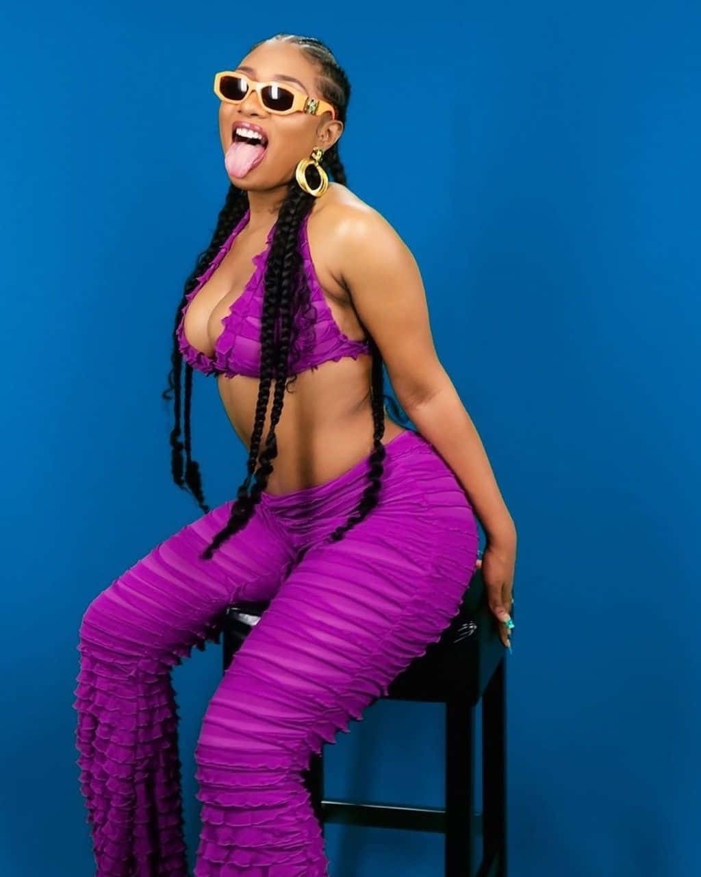 Megan Thee Stallion 桌布 壁紙 — 一位身穿鮮豔紫色皺褶服裝和黃色太陽眼鏡的女性，在純藍色攝影棚背景下頑皮地伸出舌頭。