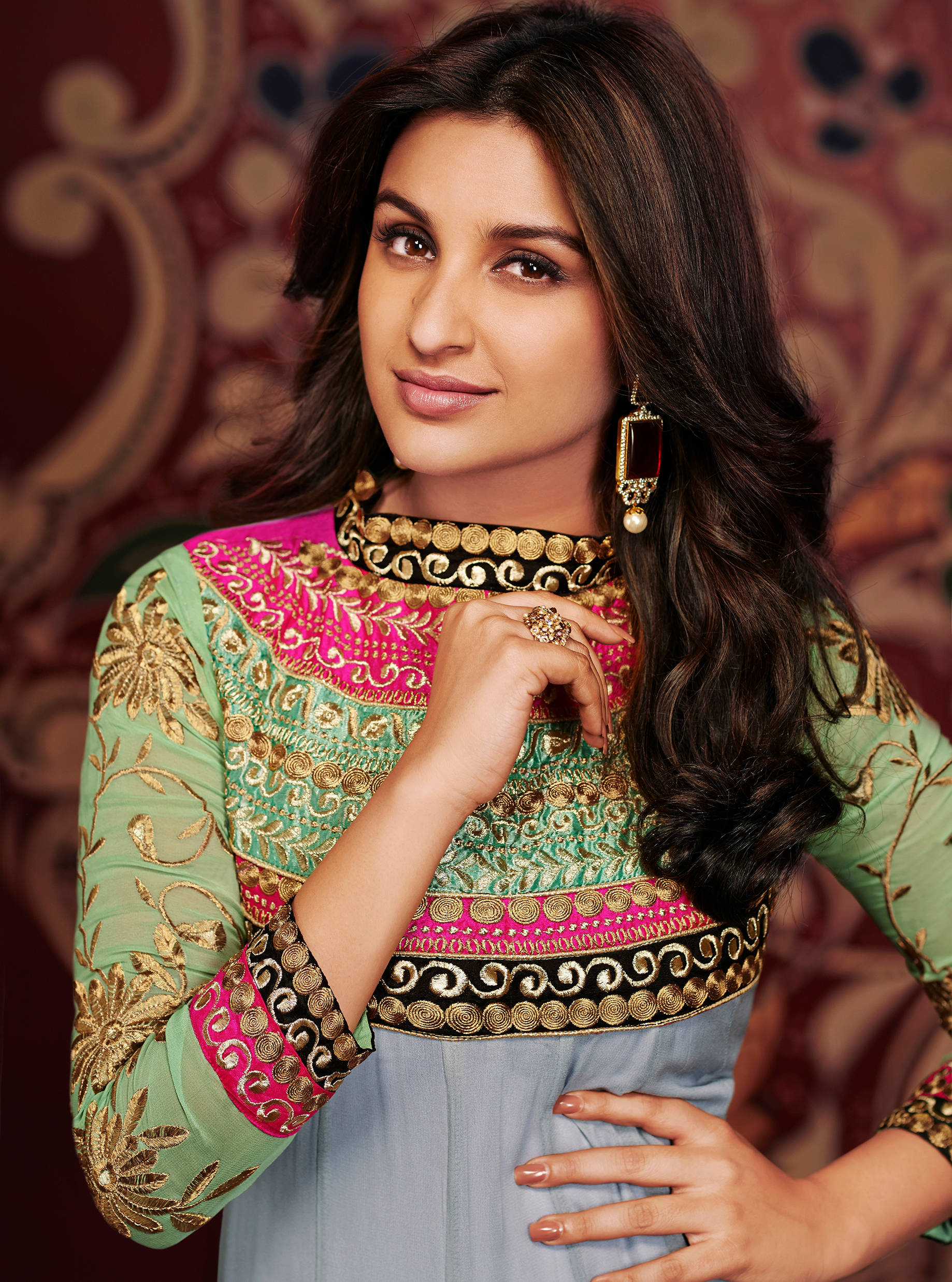 Parineeti Chopra 桌布 壁紙 — 一位身穿鮮豔綠色和粉紅色刺繡服裝的女性，在華麗圖案的紅色背景前優雅地擺姿勢。