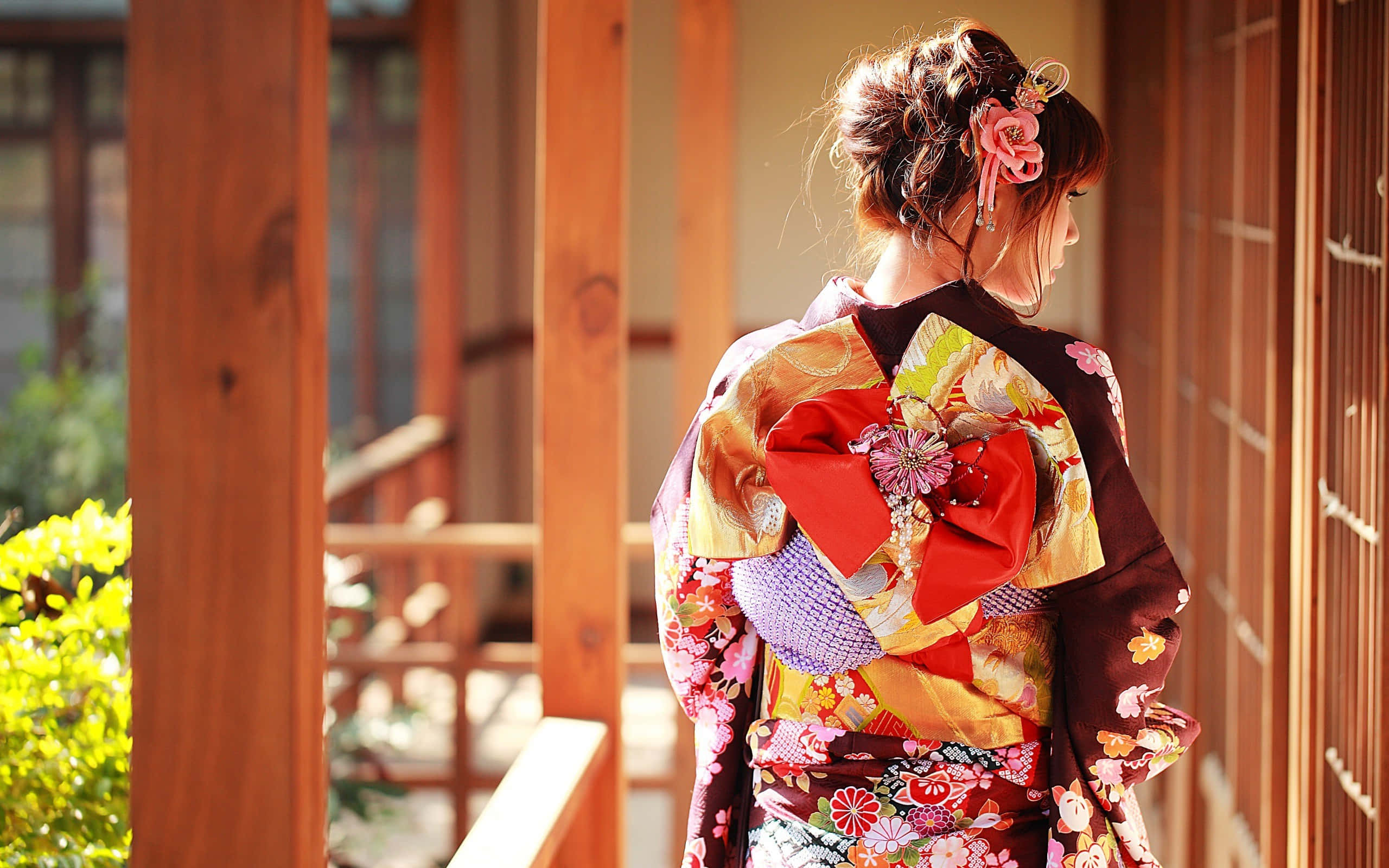 Japanese Women 桌布 壁紙 — 一位身穿鮮豔花卉和服、繫著大紅色腰帶的女性，站在沐浴在溫暖陽光下的木製陽台上。