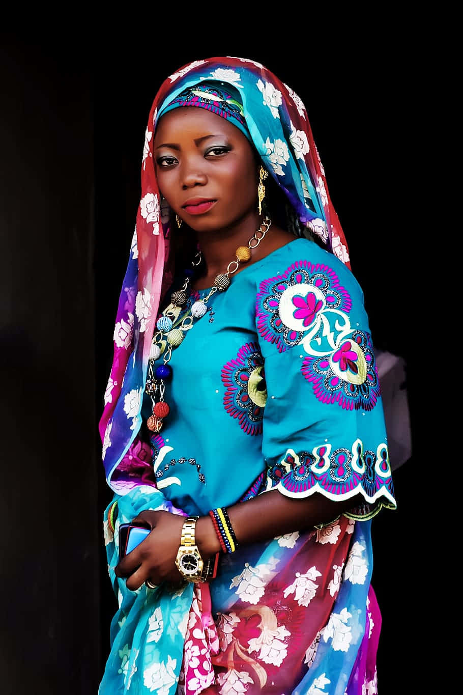Nigeria 桌布 壁紙 — 一位身穿鮮豔藍色和粉紅色花卉服飾、佩戴大膽珠寶的女性，在深色背景下自信地擺姿勢。