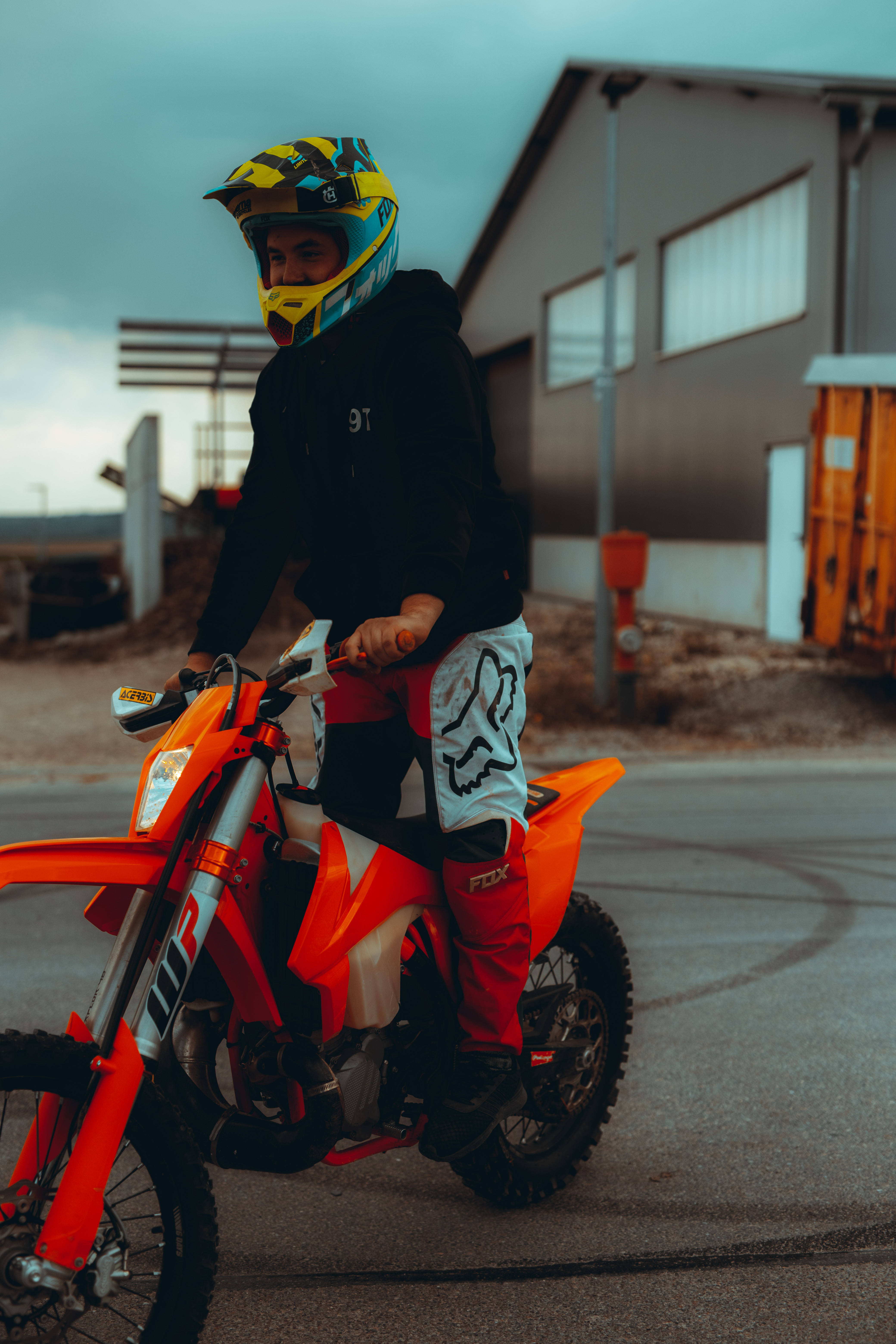 Ktm Bike 桌布 壁紙 — 一位身穿鮮豔裝備的越野摩托車手，坐在亮橙色越野車上，背景是低調的工業環境。