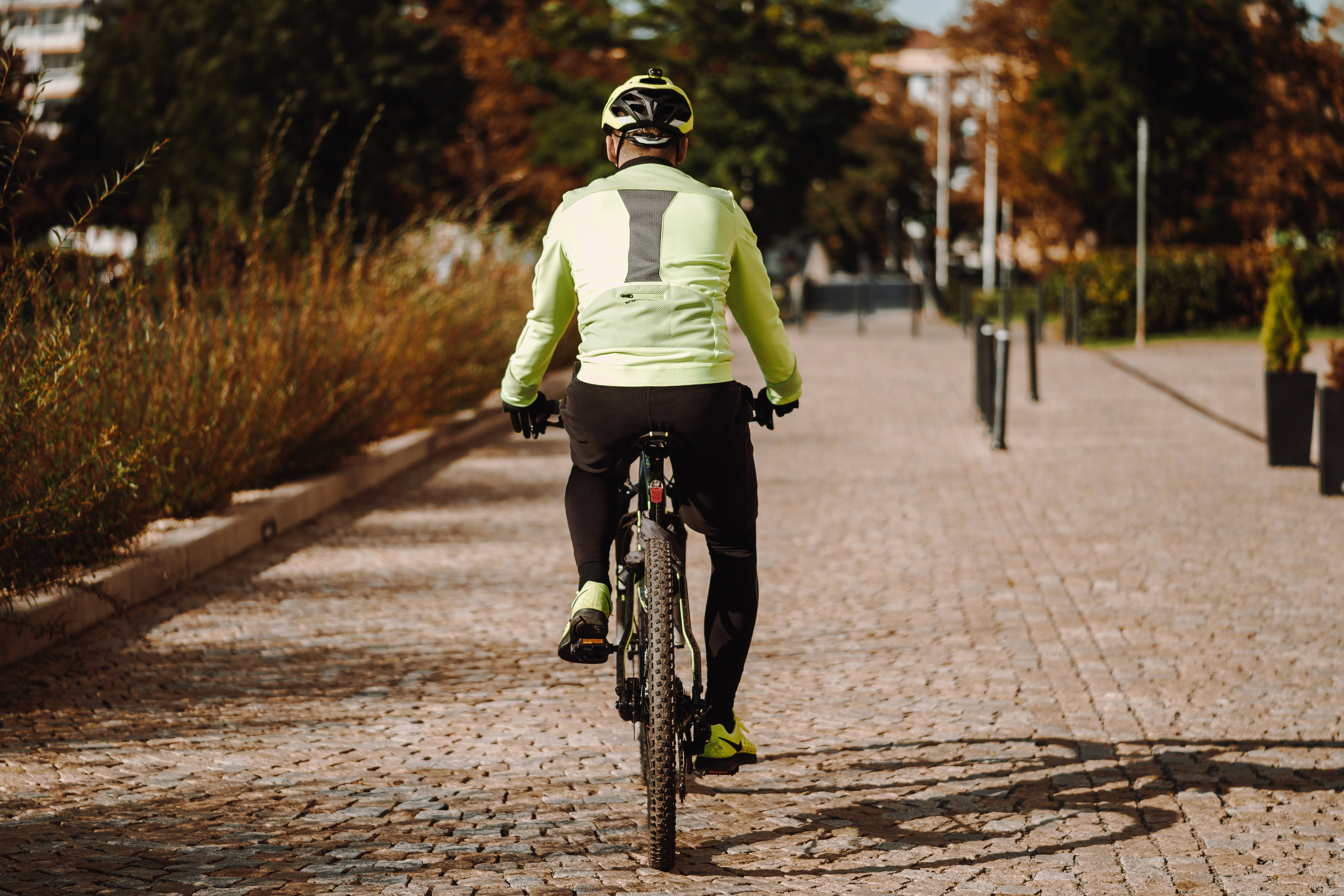 Bicycle 桌布 壁紙 — 一位身穿鮮豔霓虹裝備的自行車手，在溫暖陽光下，沿著鋪滿秋葉的鵝卵石小徑踩踏離去。