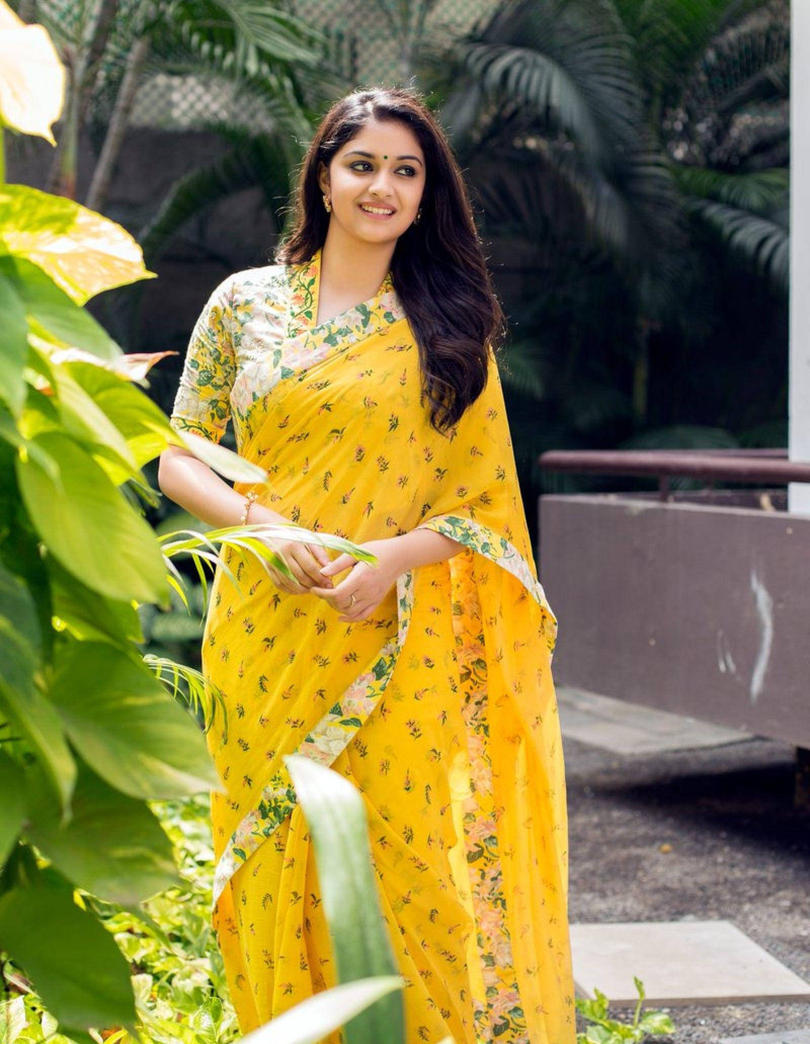 Keerthi Suresh HD 桌布 壁紙 — 一位身穿鮮豔黃色花卉紗麗的女性在明亮的日光下，於茂密的綠葉中溫柔微笑。