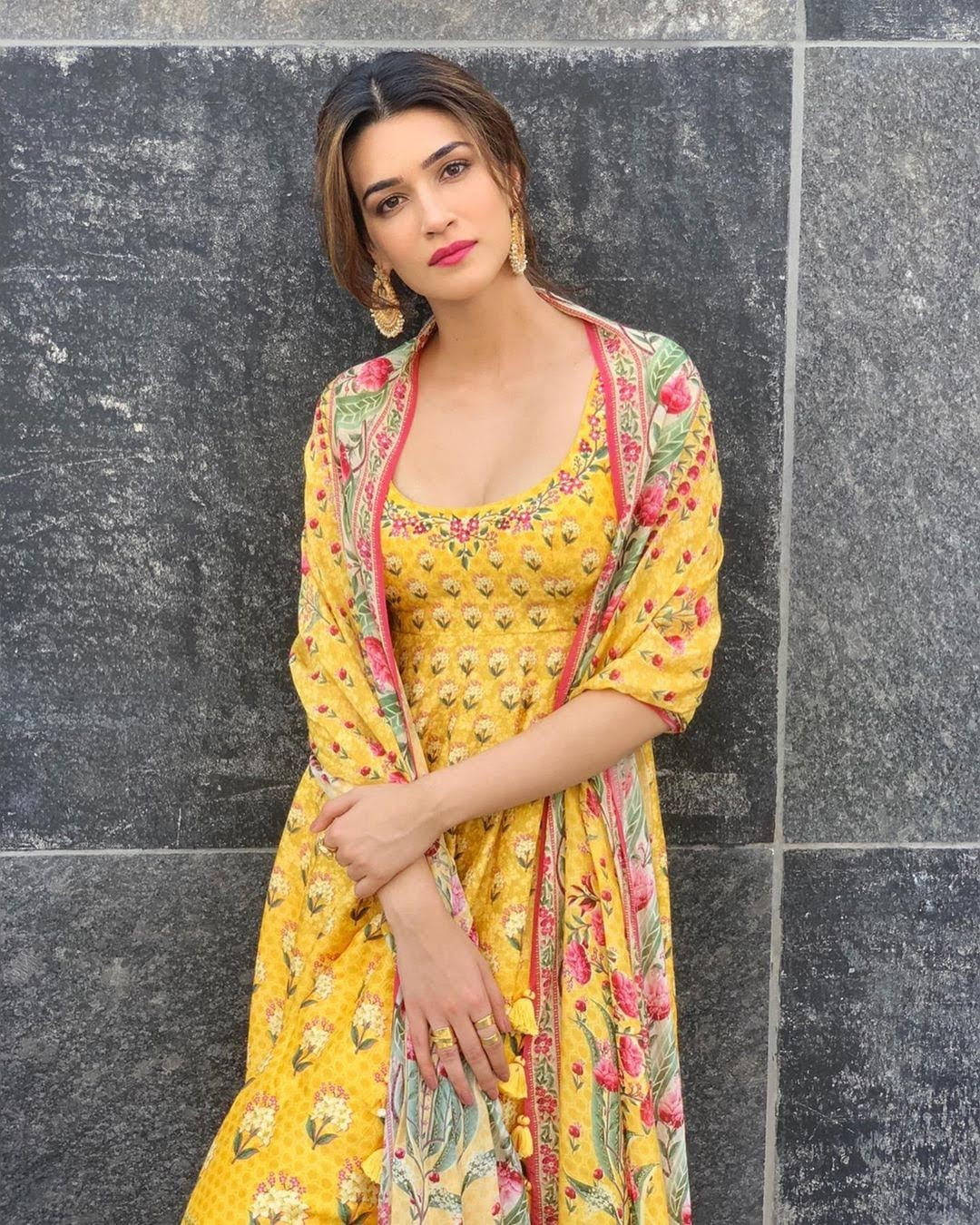 Telugu Heroines 桌布 壁紙 — 一位身穿鮮豔黃色花卉連衣裙的女性自信地站在深灰色瓷磚牆前。
