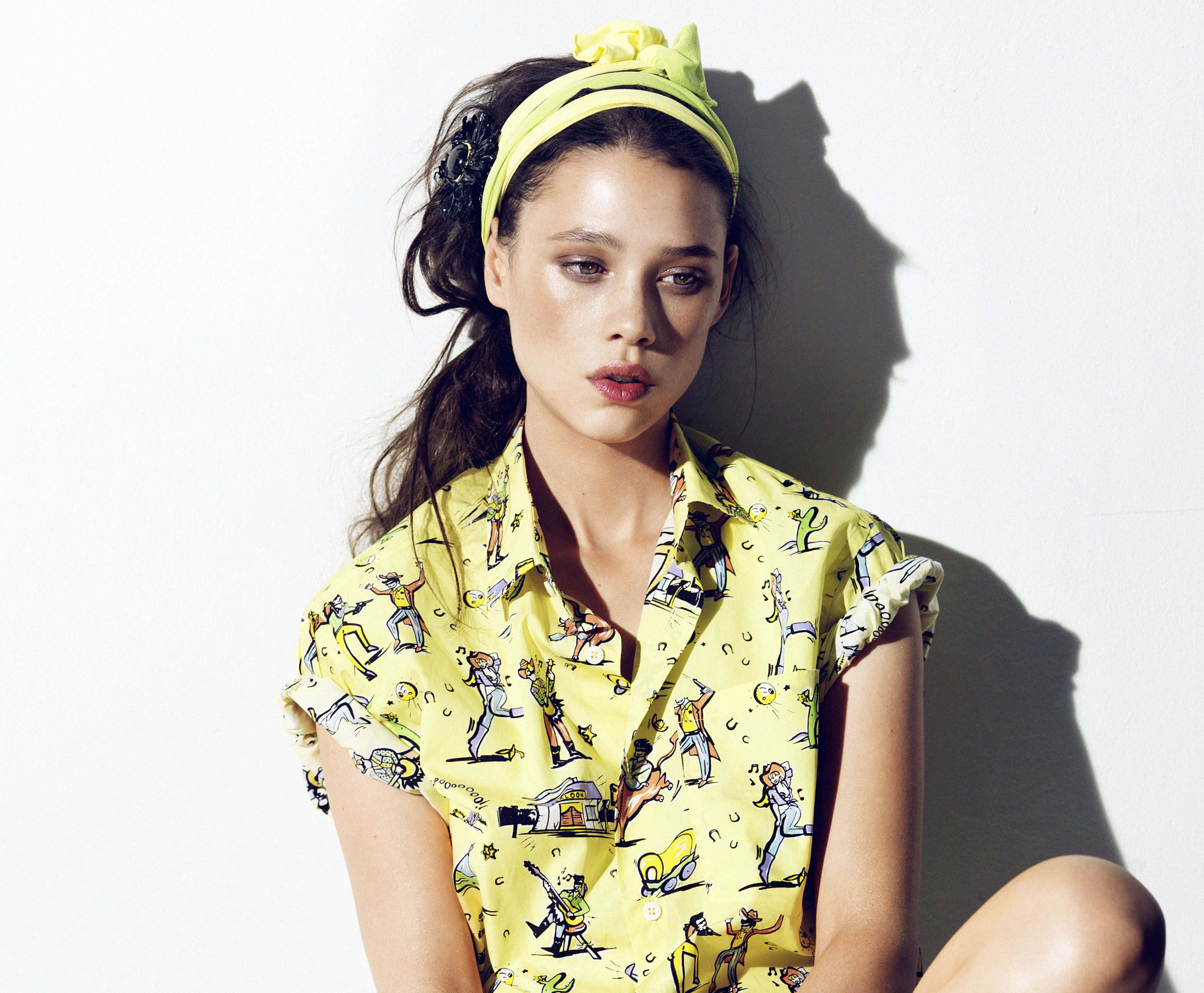 Astrid Berges Frisbey 桌布 壁紙 — 一位身穿鮮黃色圖案襯衫和相配頭帶的女性，在純白牆壁前擺出姿勢，投射出戲劇性的陰影。