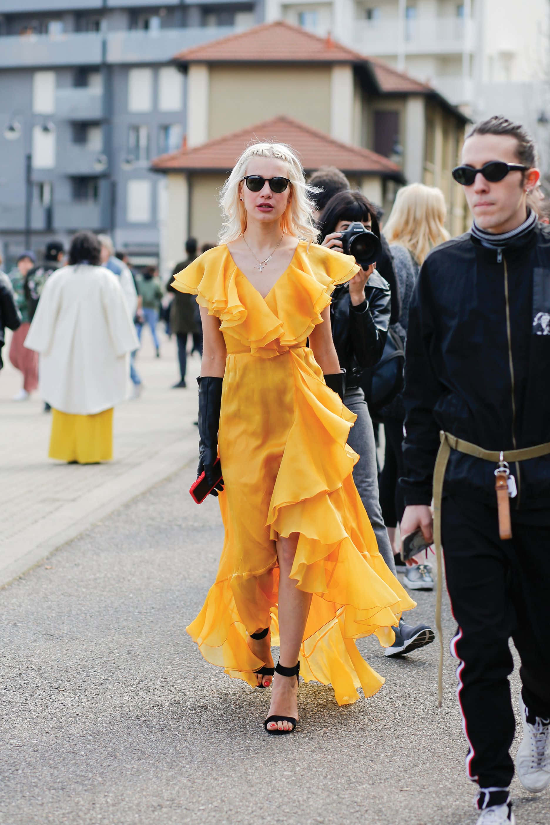 Yellow Fashion 桌布 壁紙 — 一位身穿鮮黃色荷葉邊連衣裙和黑色手套的女性，自信地走在滿是行人的城市街道上。