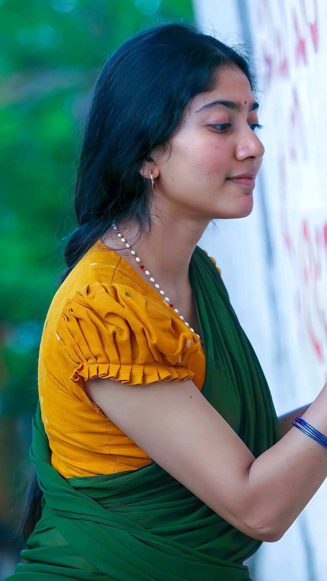 Sai Pallavi 桌布 壁紙 — 一位身穿鮮黃色襯衫和綠色紗麗的女性，在模糊的綠色和白色背景下若有所思地望向一旁。