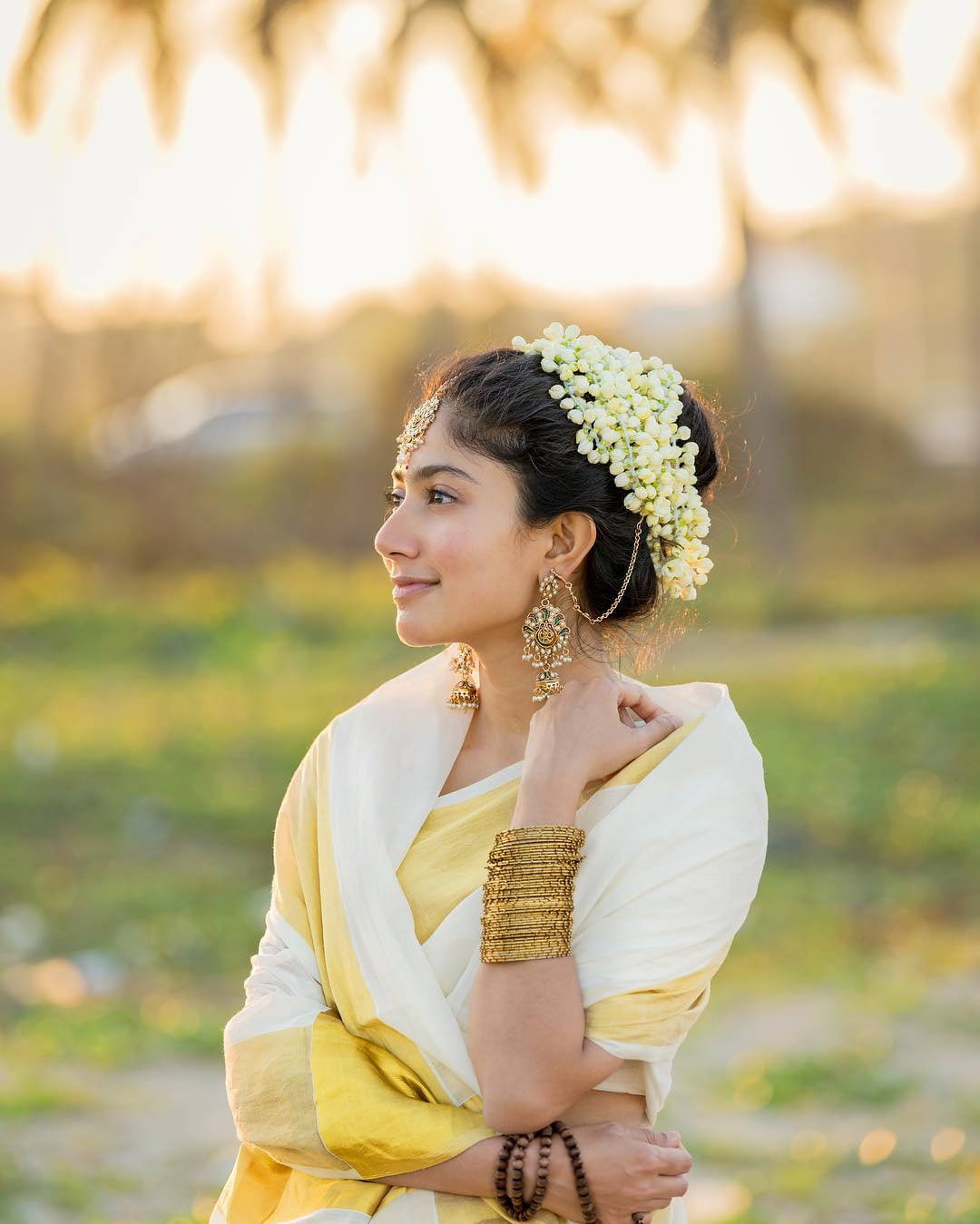 Sai Pallavi 桌布 壁紙 — 一位身穿黃白傳統服飾、飾有花卉頭飾和金飾的女子，若有所思地凝視著日落。