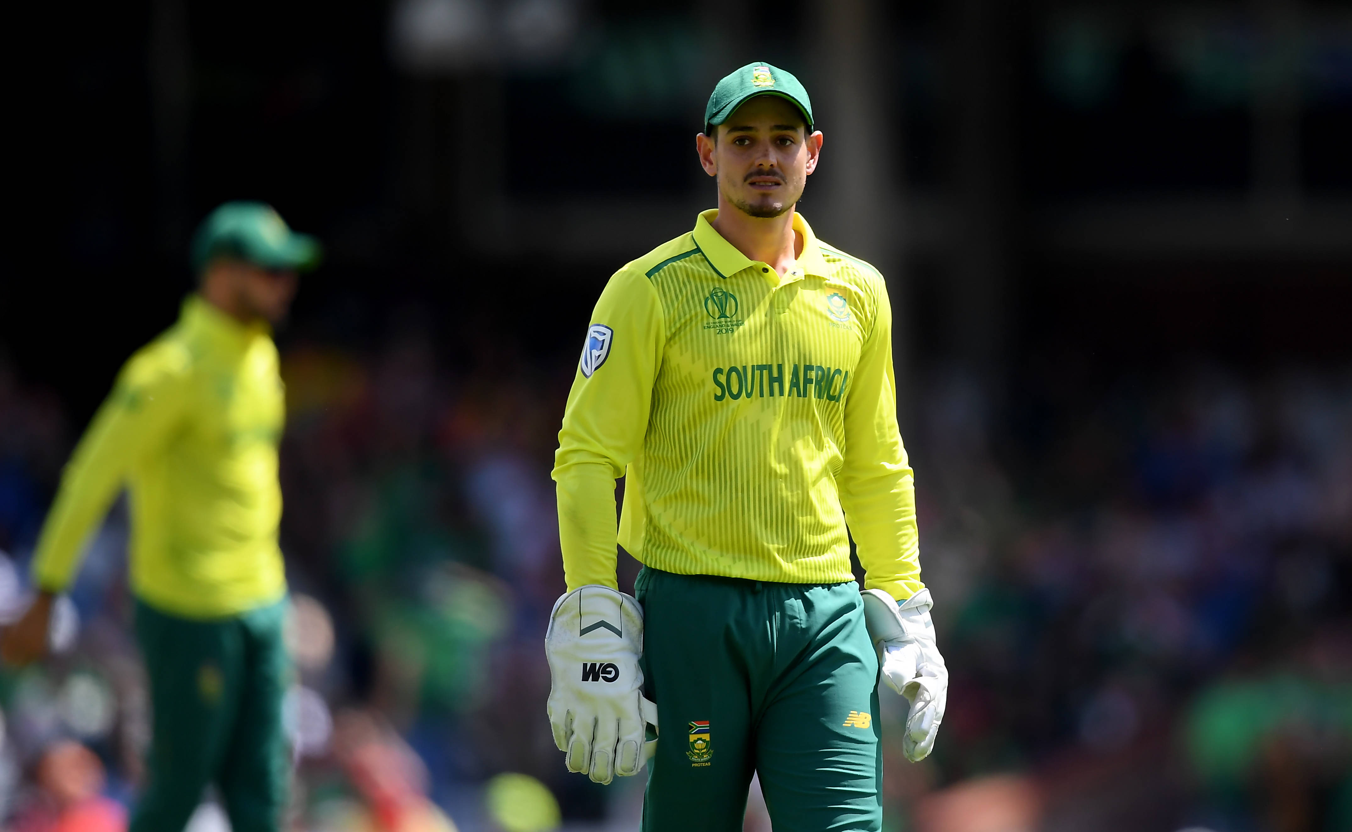quinton de kock 桌布 壁紙 — 一位身穿黃綠色球衣的南非板球運動員在陽光明媚的球場上莊嚴地行走，背景是模糊的觀眾。