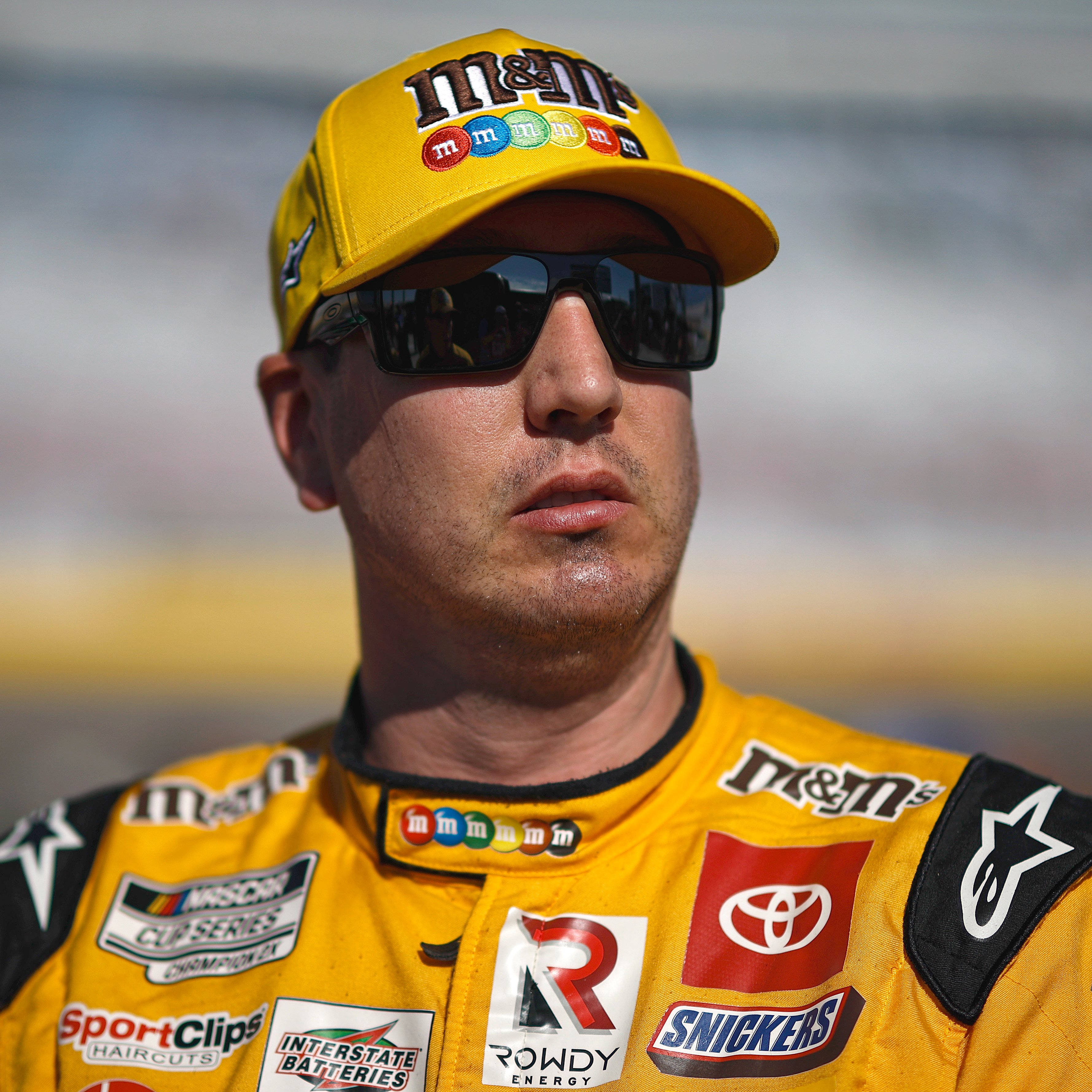 運動 桌布 壁紙 — 一位身穿黃色M&M's賽車服和帽子的NASCAR車手，在太陽眼鏡下專注地凝視著陽光照耀的賽道。