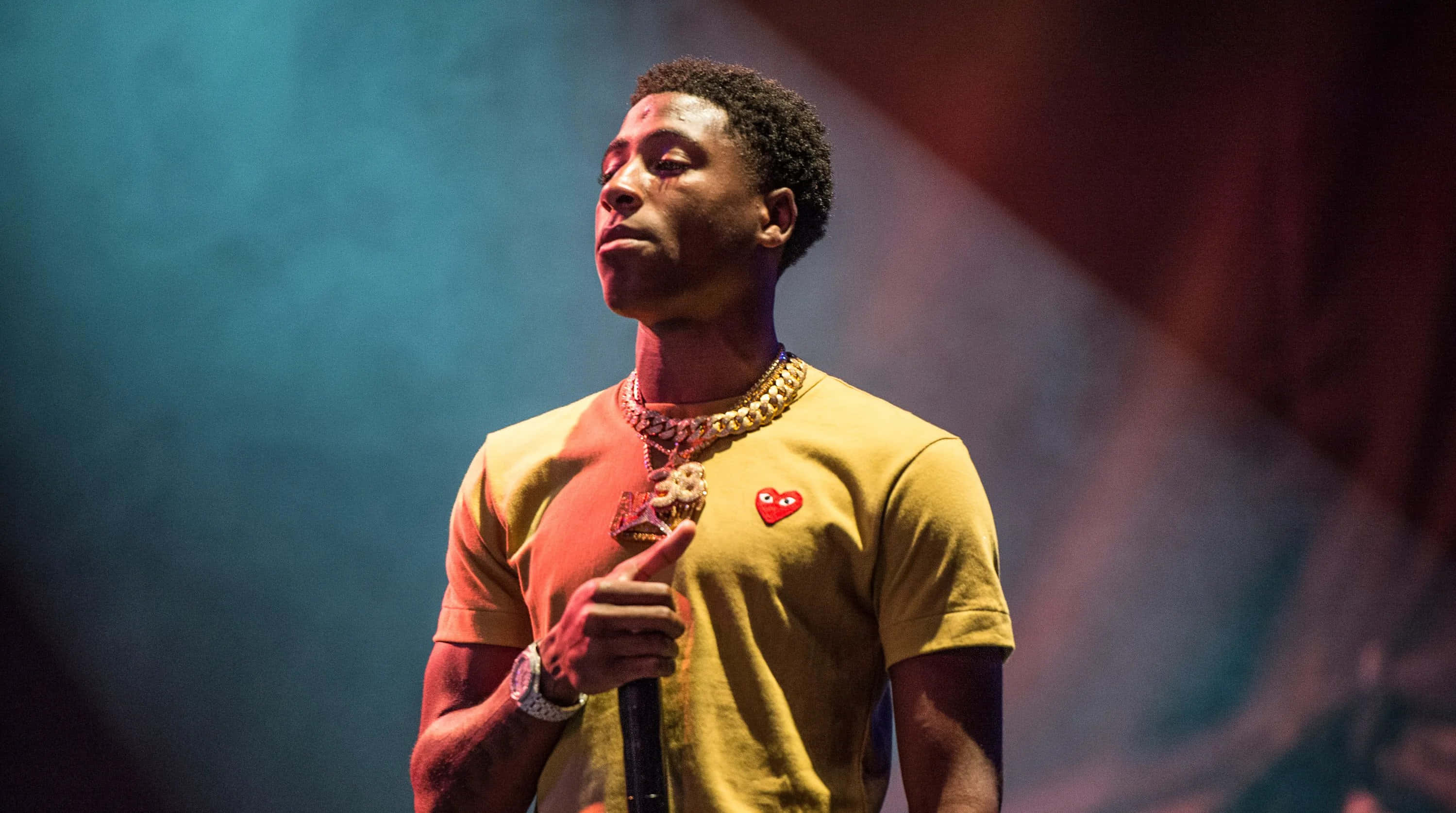 NBA YoungBoy 桌布 壁紙 — 一位身穿黃色T恤和金色鏈條的男性表演者在戲劇性的藍色和紅色舞台燈光下拿著麥克風。