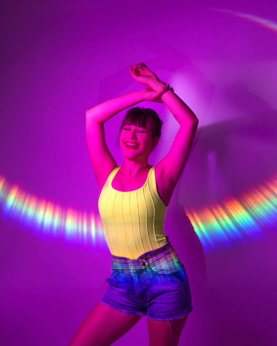 Photoshoot 桌布 壁紙 — 一位身穿黃色上衣和牛仔短褲的快樂女性，在背後有彩虹弧線的鮮豔紫色燈光下跳舞。