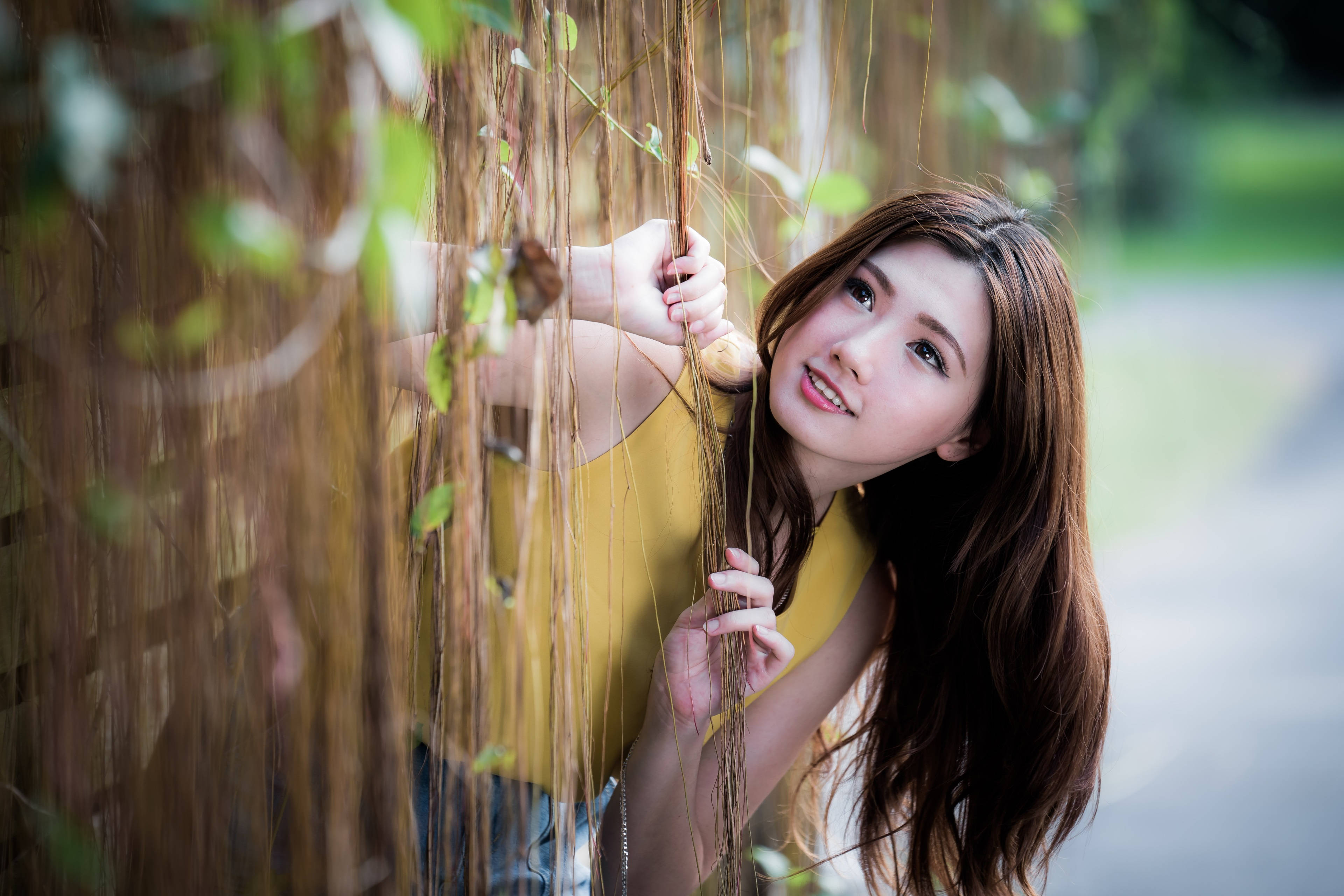 美女 桌布 壁紙 — 一位身穿黃色上衣的年輕女子在陽光明媚的花園中，透過懸掛的藤蔓露出溫柔的微笑。