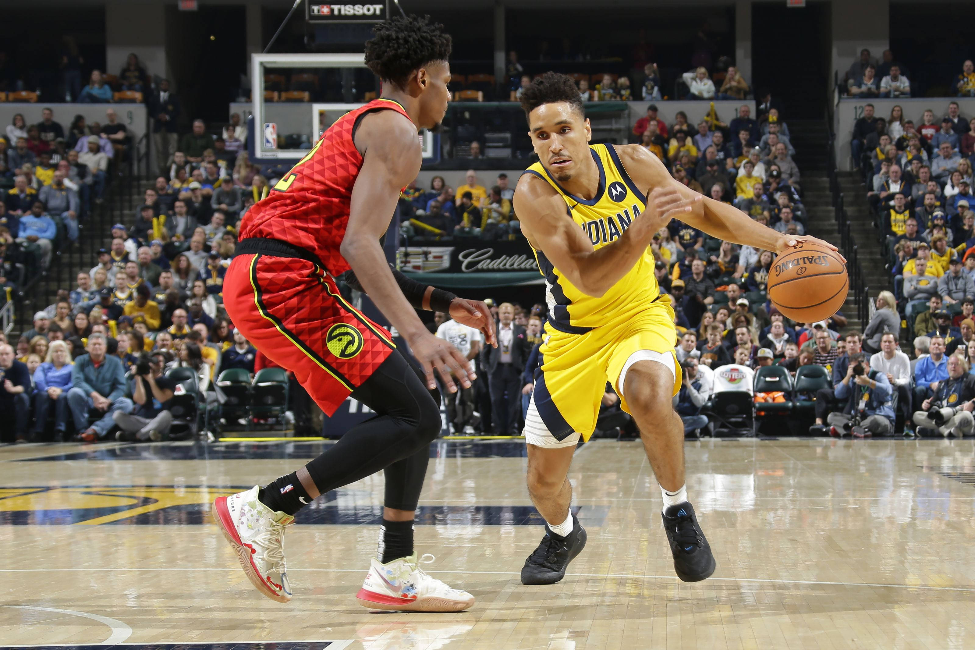 malcolm brogdon 桌布 壁紙 — 一位身穿黃色印第安納球衣的籃球運動員在擠滿觀眾的場館比賽中運球，同時被一位身穿紅色多倫多球衣的對手防守。