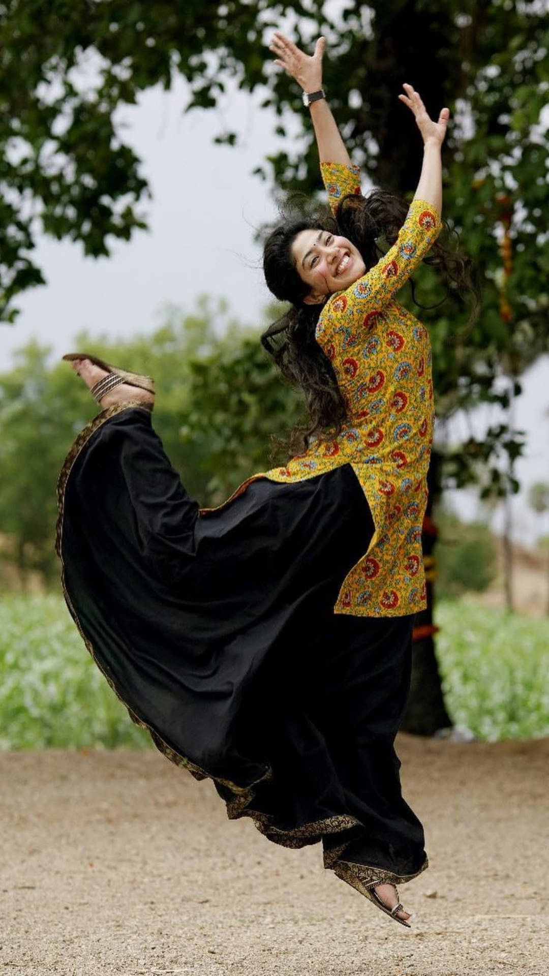 Sai Pallavi 桌布 壁紙 — 一位身穿黃色圖案上衣和黑色飄逸長褲的快樂女性，在枝葉茂密的樹下雙臂高舉跳躍在空中。