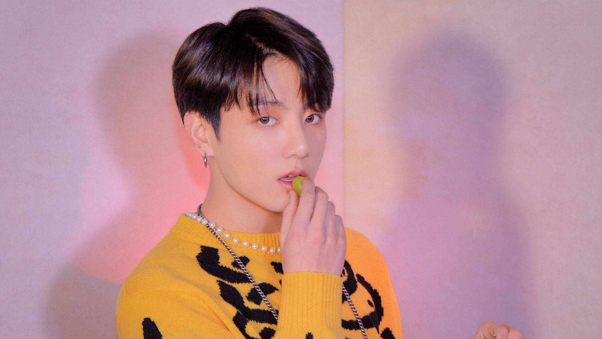 Jungkook 桌布 壁紙 — 一位身穿黃色圖案毛衣的年輕男子咬著一顆青葡萄，同時看著鏡頭，背景是柔和的粉紅色和紫色燈光牆壁。