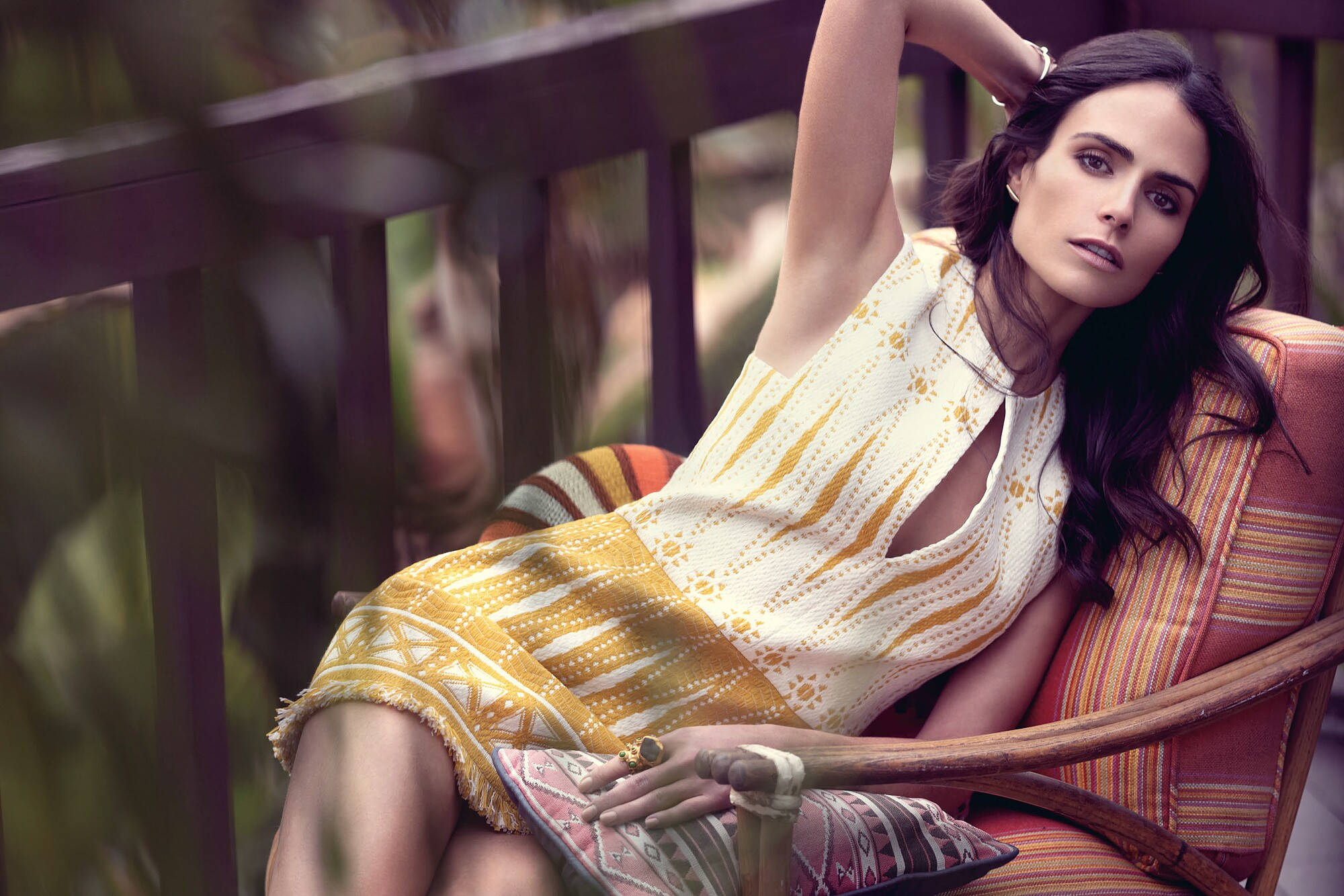 Jordana Brewster 桌布 壁紙 — 一位身穿黃色圖案連衣裙的女性優雅地躺在被綠植環繞的門廊木椅上。