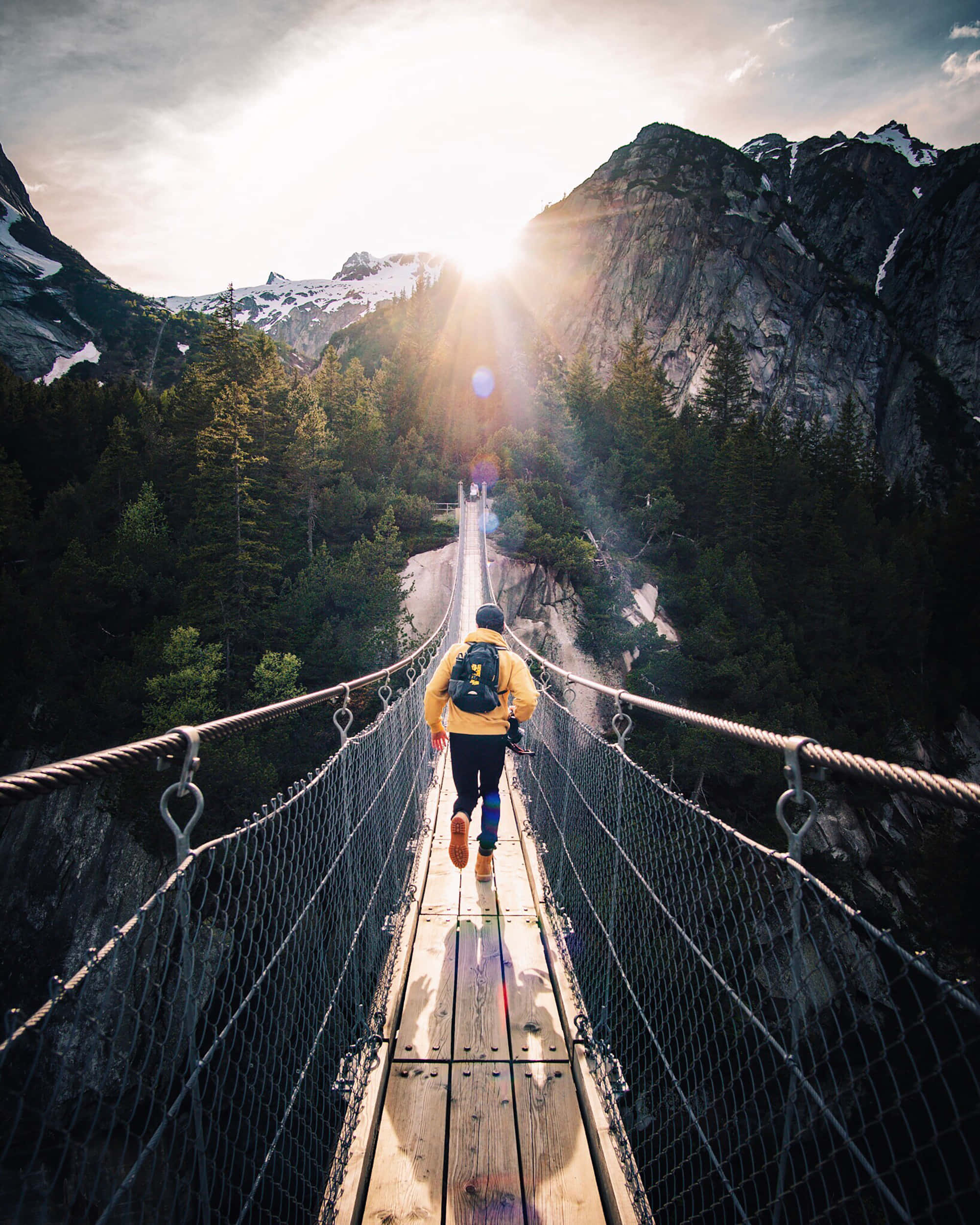bridge 桌布 壁紙 — 一位身穿黃色外套的跑者穿過木製吊橋，金色陽光在積雪的山峰之間迸發。