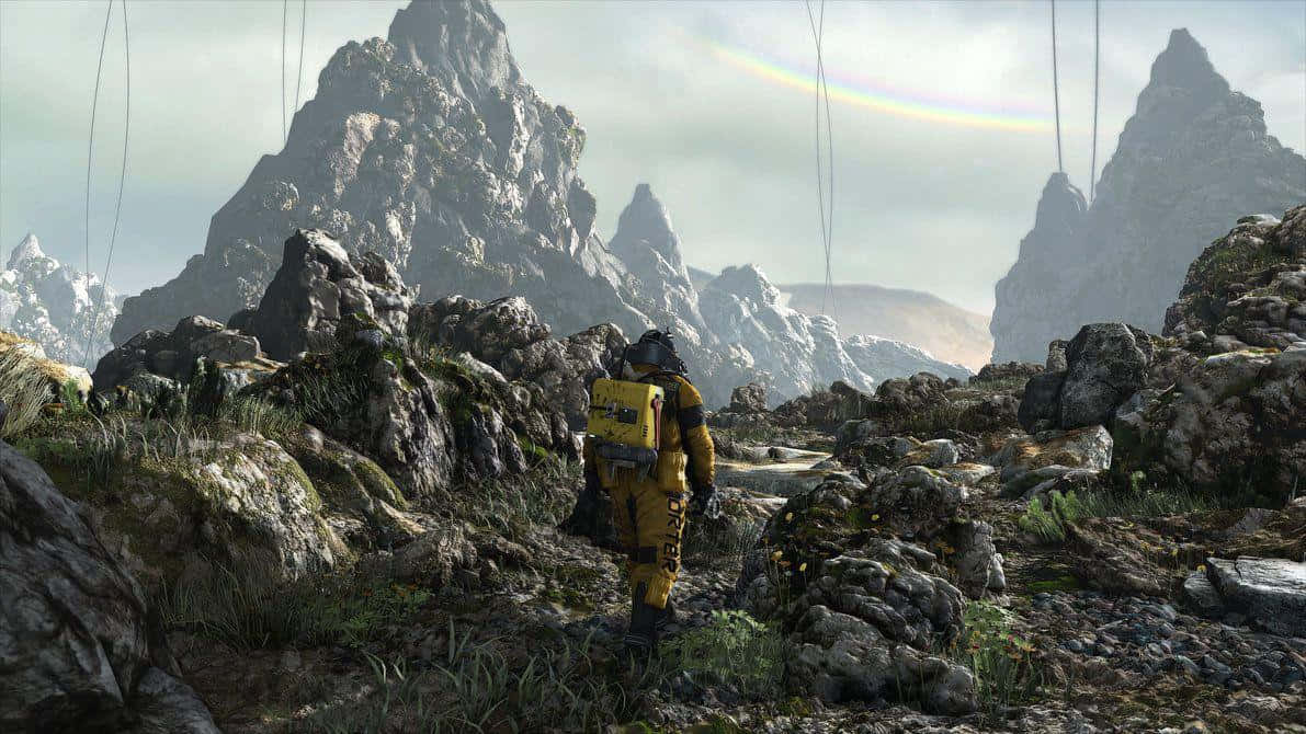 Death Stranding Pc 桌布 壁紙 — 一位身穿黃色套裝的孤獨探險家，在微弱的彩虹下走過岩石山隘。