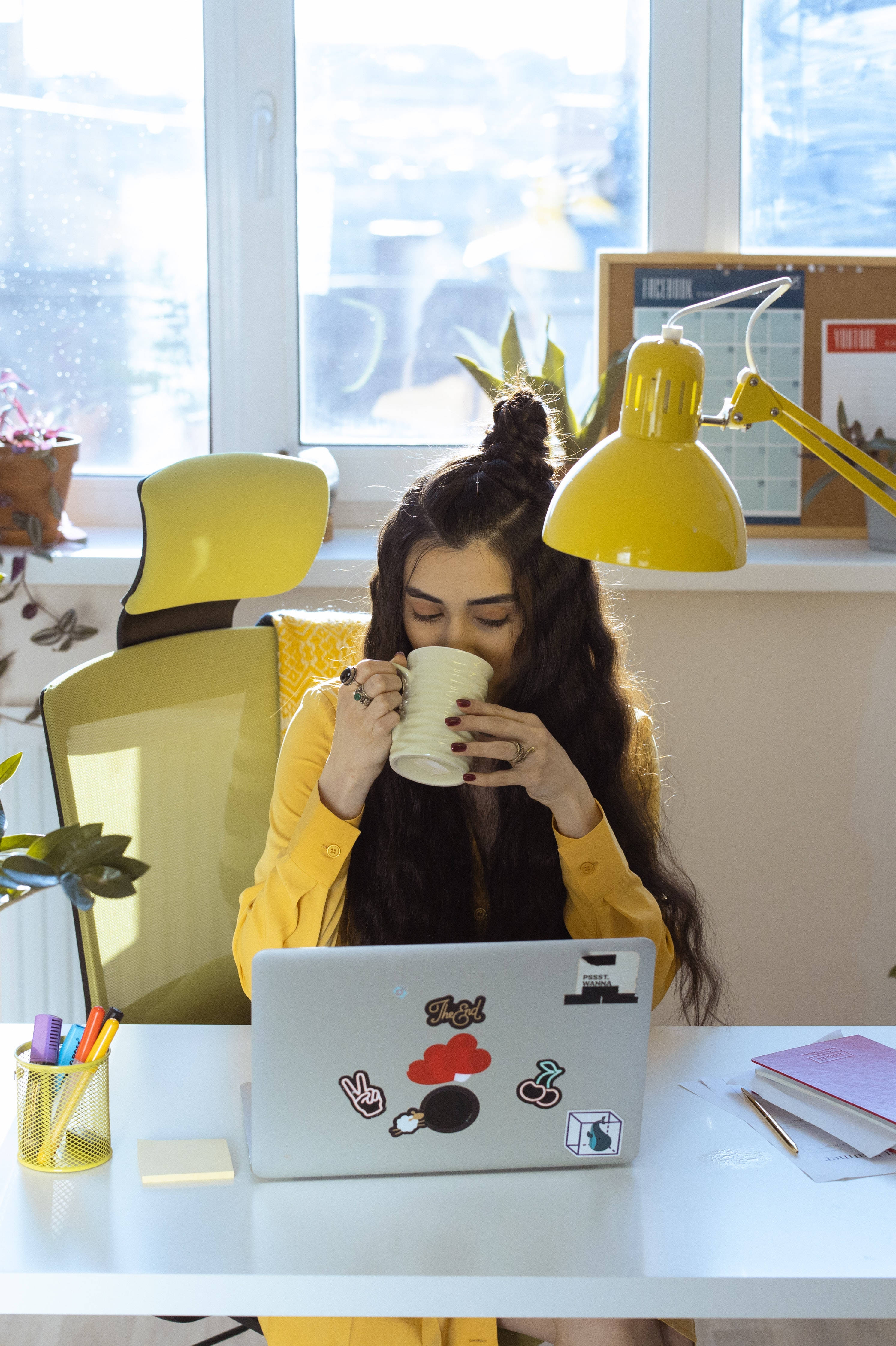 aesthetic tumblr laptop 桌布 壁紙 — 一位身穿黃色服裝的年輕女性在陽光照射的書桌前，拿著馬克杯啜飲，桌上有一部筆記型電腦和一盞黃色檯燈。