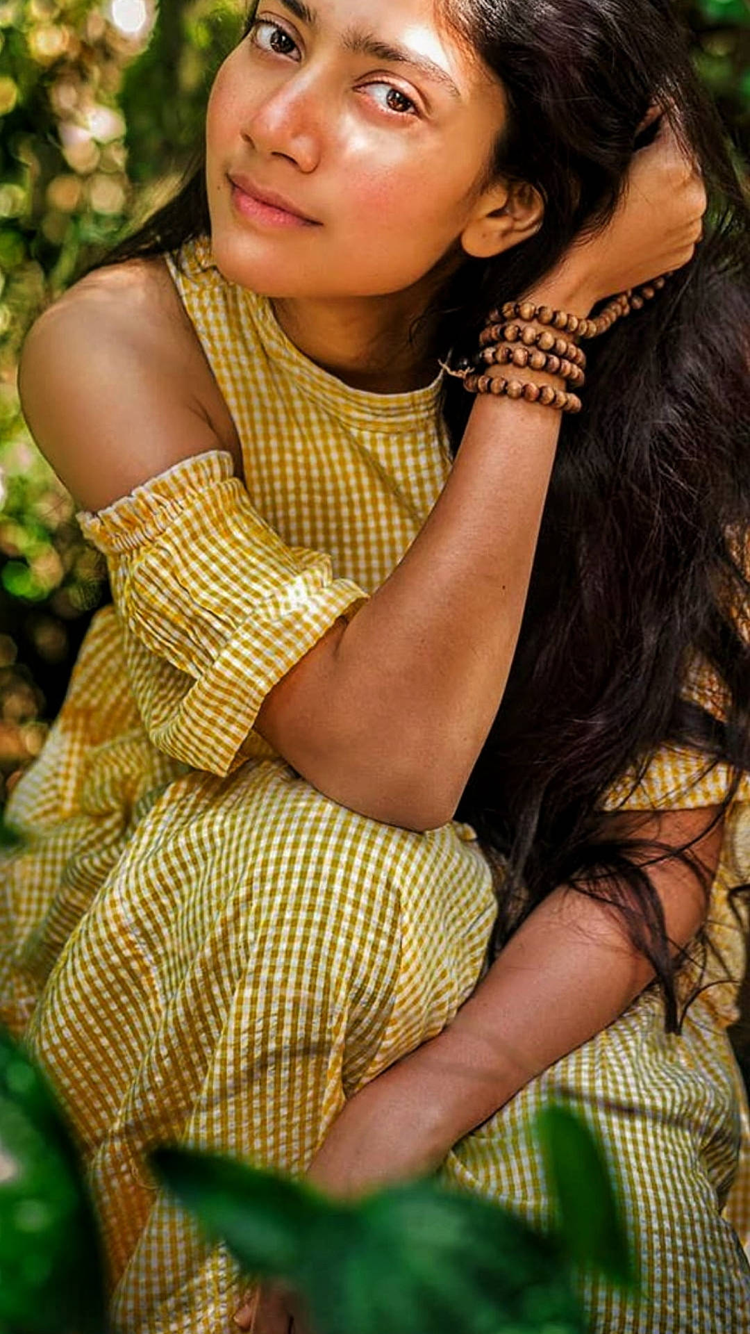 Sai Pallavi 桌布 壁紙 — 一位身穿黃色格子裙的年輕女性在茂密的綠葉中輕撫頭髮，溫柔微笑。