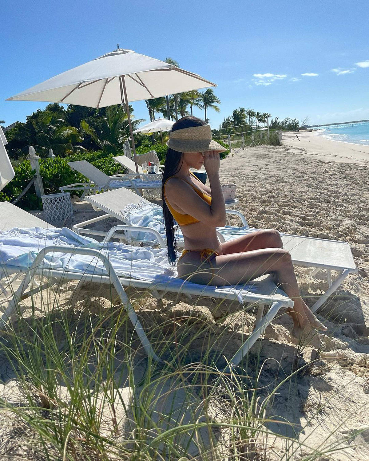 Kourtney 桌布 壁紙 — 一位身穿黃色比基尼和草帽遮陽板的女士，在大傘下的白色沙灘椅上休息，背景是藍綠色的海洋和棕櫚樹。