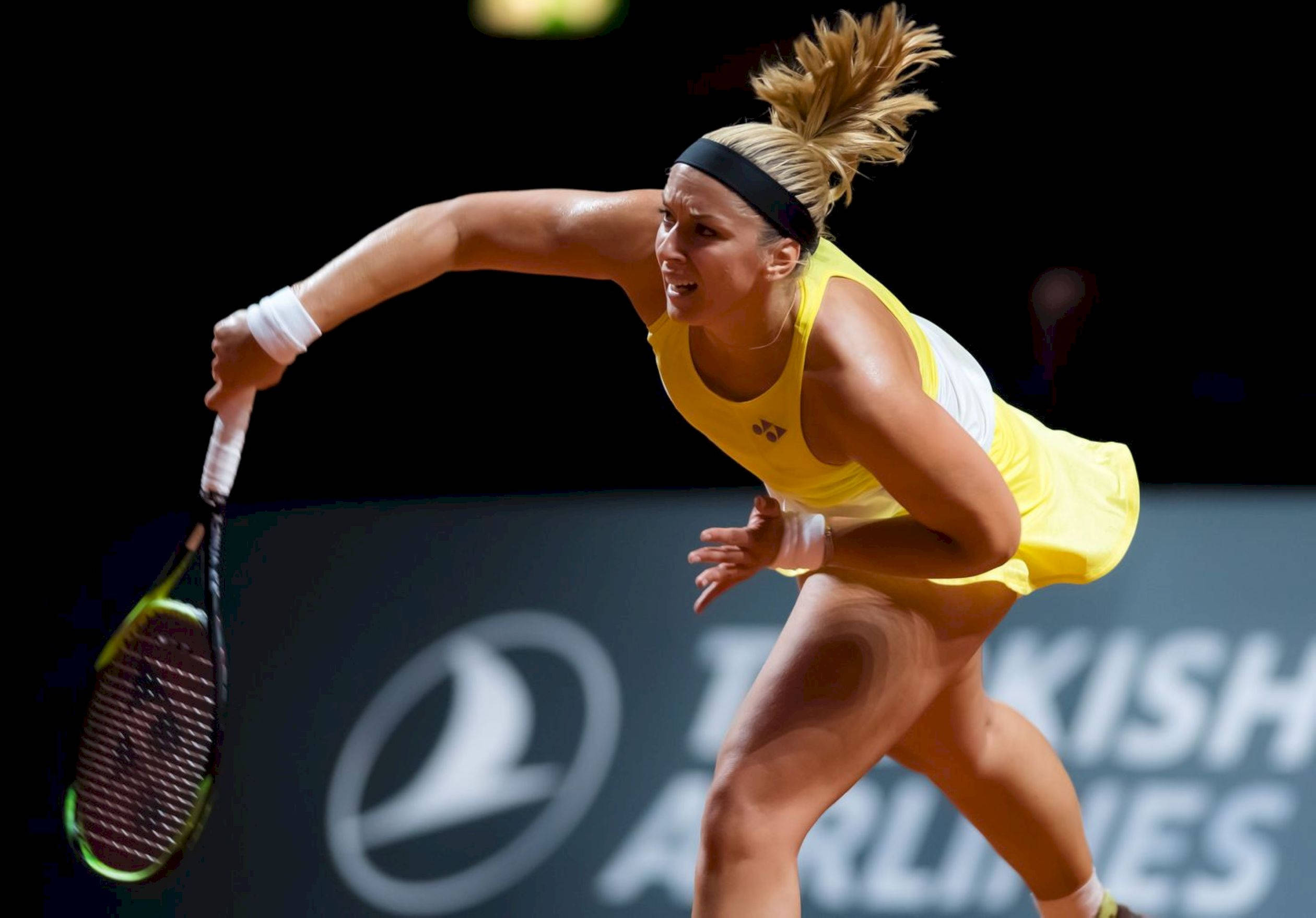 Tennis 桌布 壁紙 — 一位身穿黃色球衣的女網球選手在明亮的體育場燈光下，專注地向前猛衝，進行強力擊球。