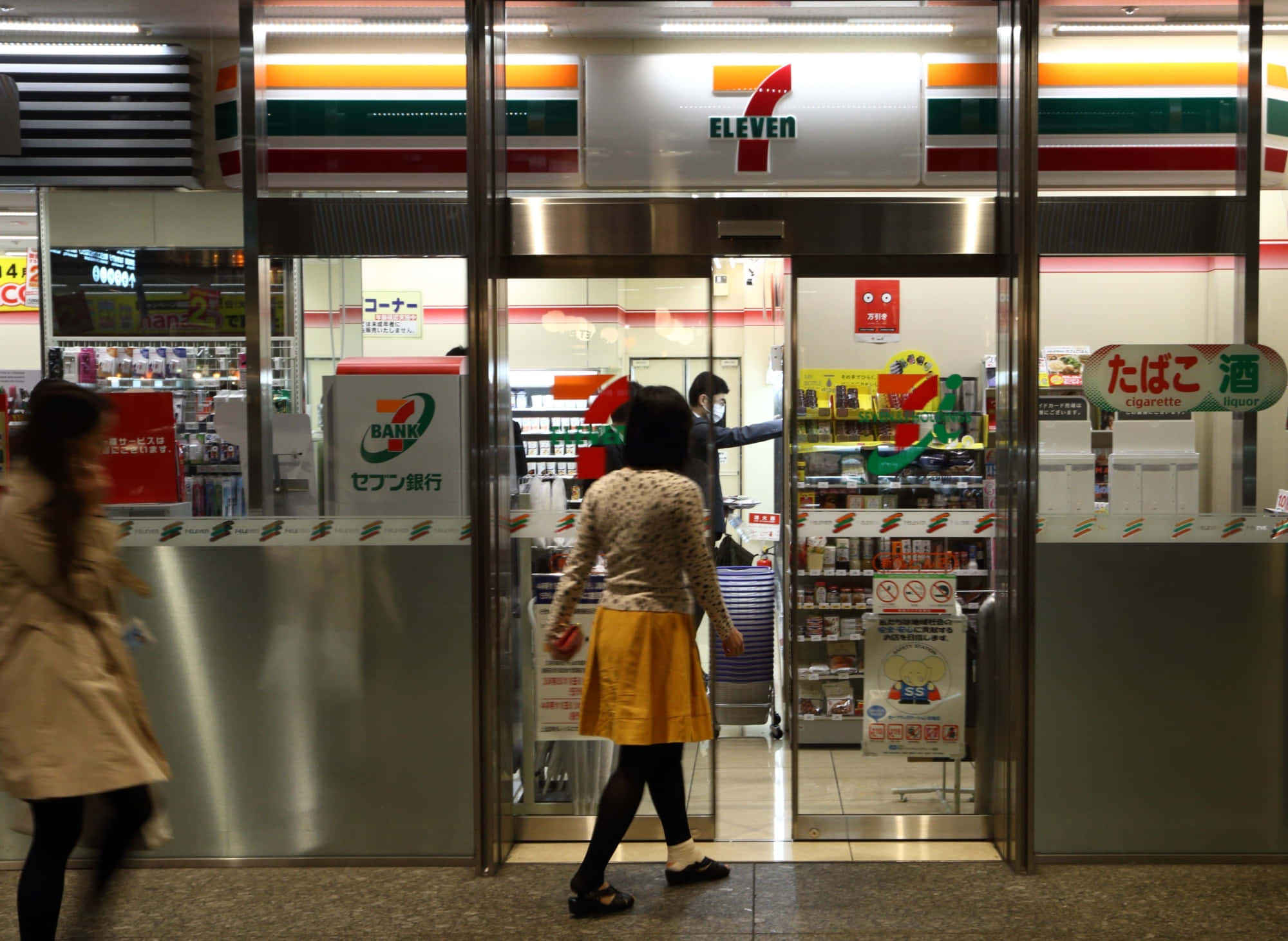 7-ELEVEN 桌布 壁紙 — 一位身穿黃色裙子的女士在夜間走進一間燈火通明的7 Eleven店鋪，店鋪設有玻璃門和霓虹招牌。