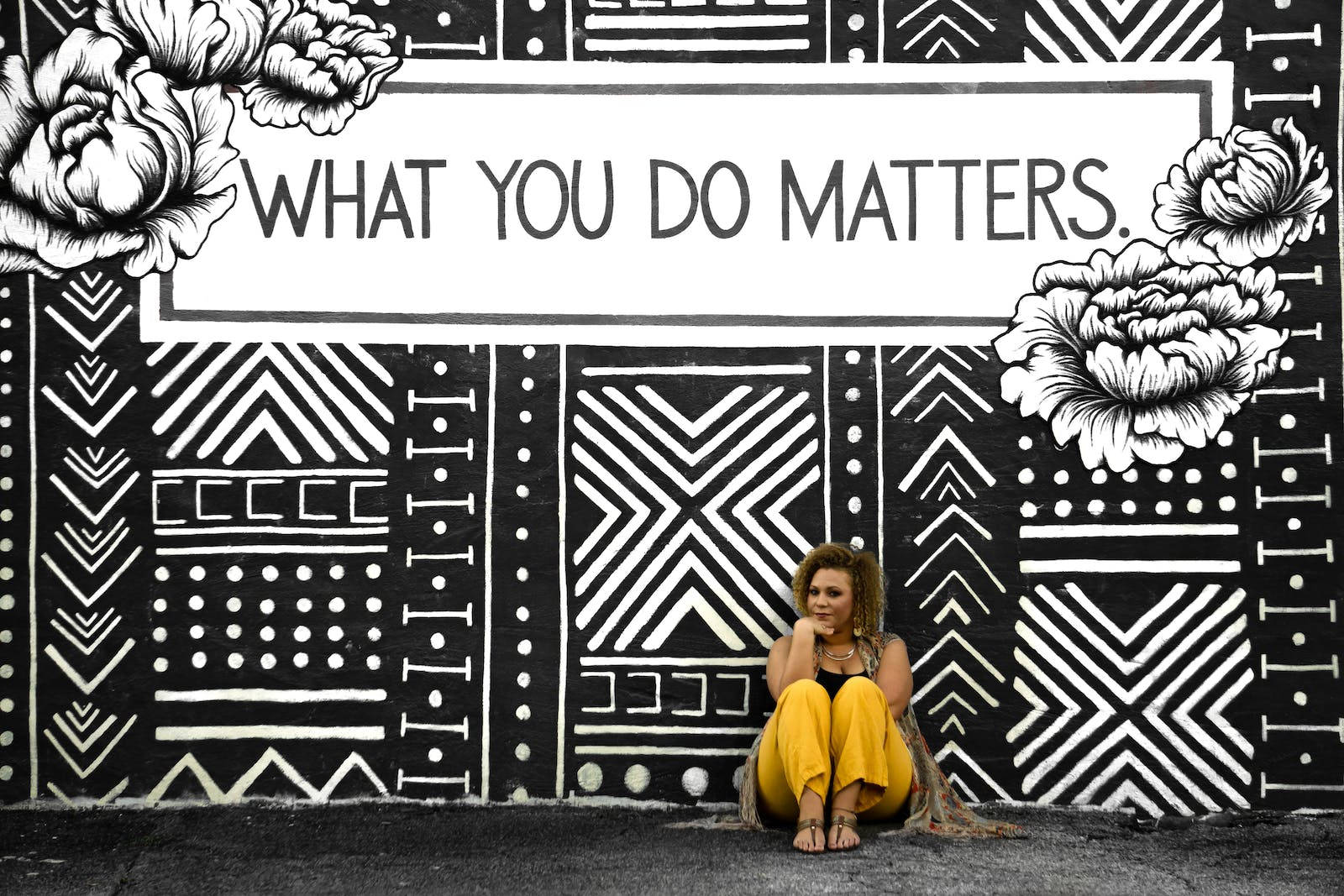 Motivation 桌布 壁紙 — 一位身穿黃色褲子的女士，若有所思地坐在一幅大膽的黑白幾何壁畫前，壁畫帶有花卉點綴和「WHAT YOU DO MATTERS」字句。