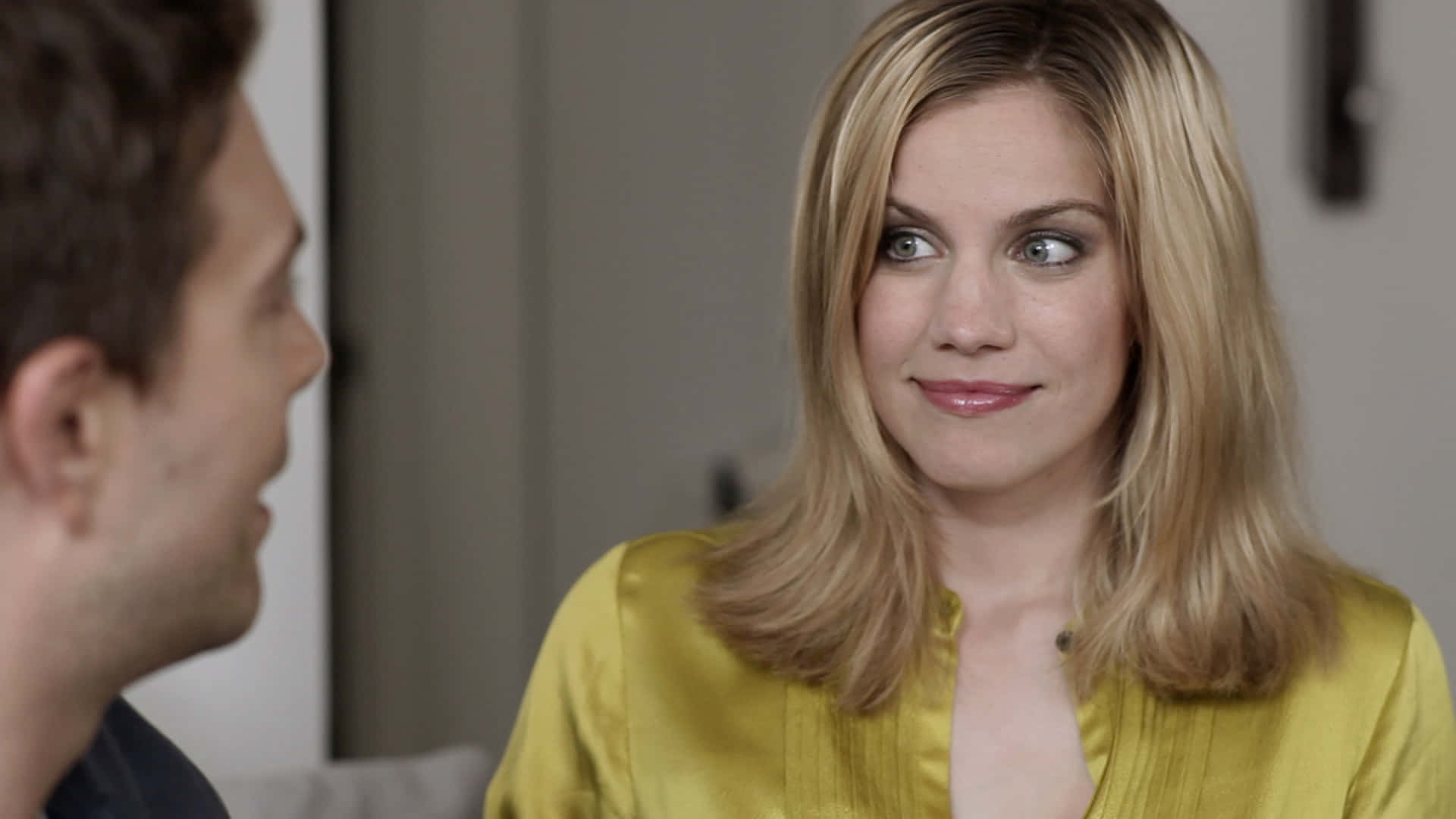 anna chlumsky 桌布 壁紙 — 一位身穿黃色襯衫的女性在柔和光線的室內環境中與一名男性交談，露出意味深長的微笑。