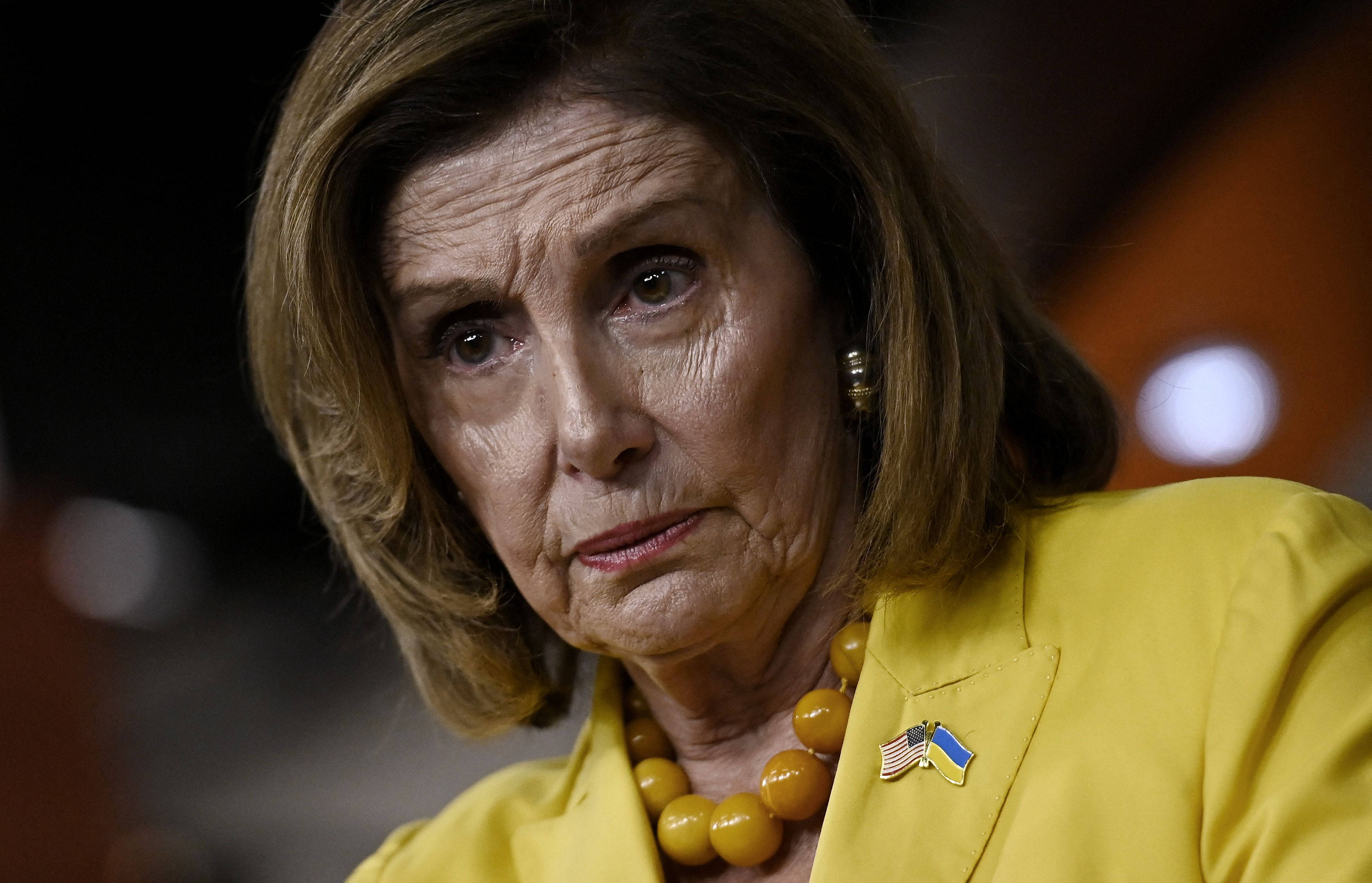 Nancy Pelosi 桌布 壁紙 — 一位身穿黃色西裝外套、戴著珠鏈和烏克蘭國旗別針的女性特寫肖像，表情嚴肅地向下看。