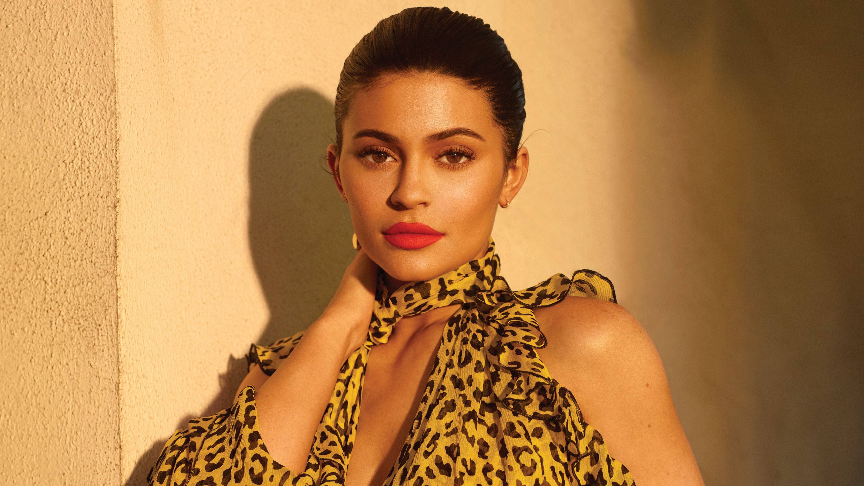 Kylie 桌布 壁紙 — 一位身穿黃色豹紋上衣的女性，在陽光照射的紋理牆前擺姿勢，擁有鮮豔的紅唇和造型的頭髮。