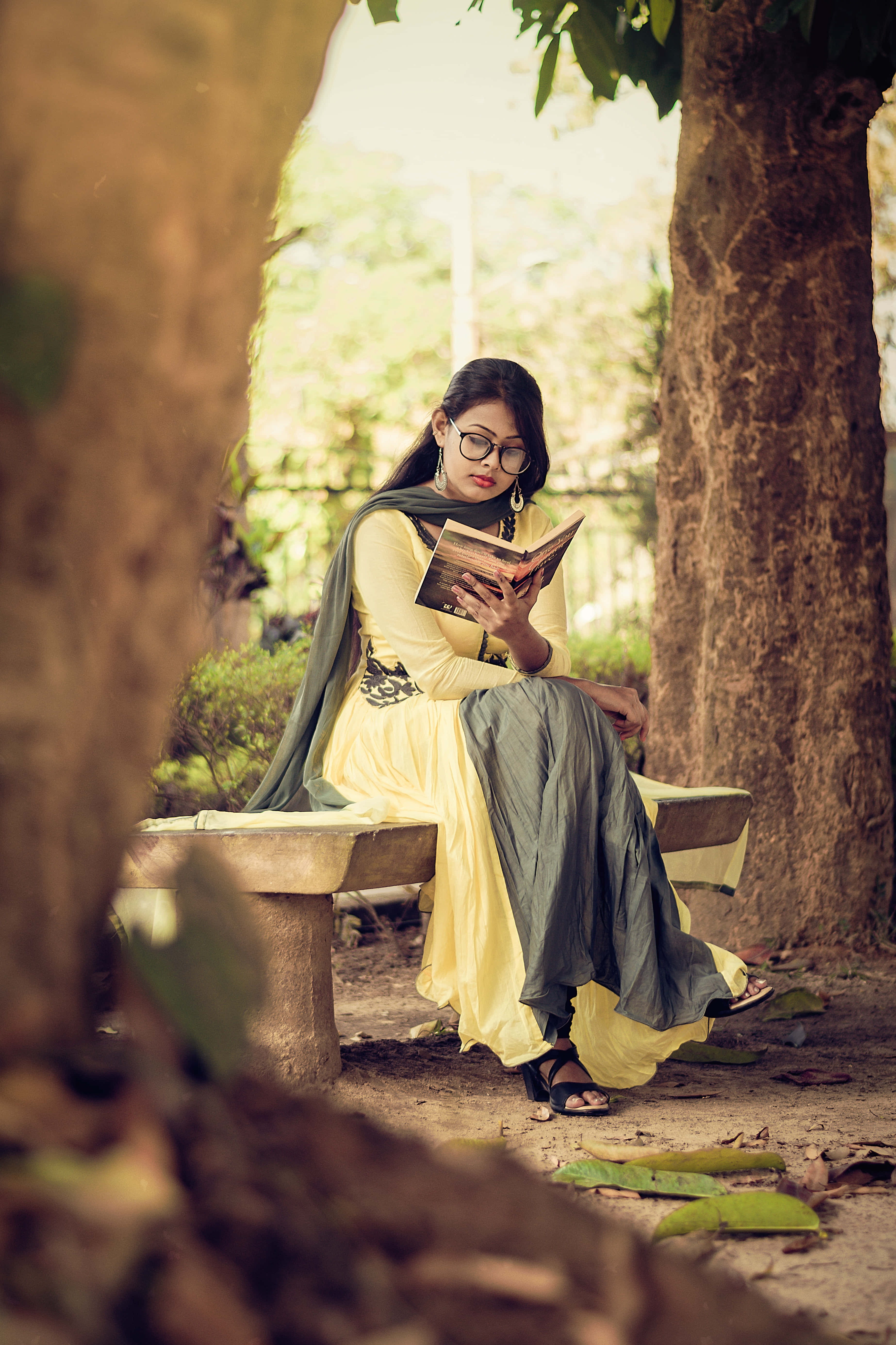 Reading 桌布 壁紙 — 一位身穿黃色連衣裙、戴著眼鏡的女性，在陽光明媚的花園裡，坐在大樹幹之間的石凳上閱讀一本書。