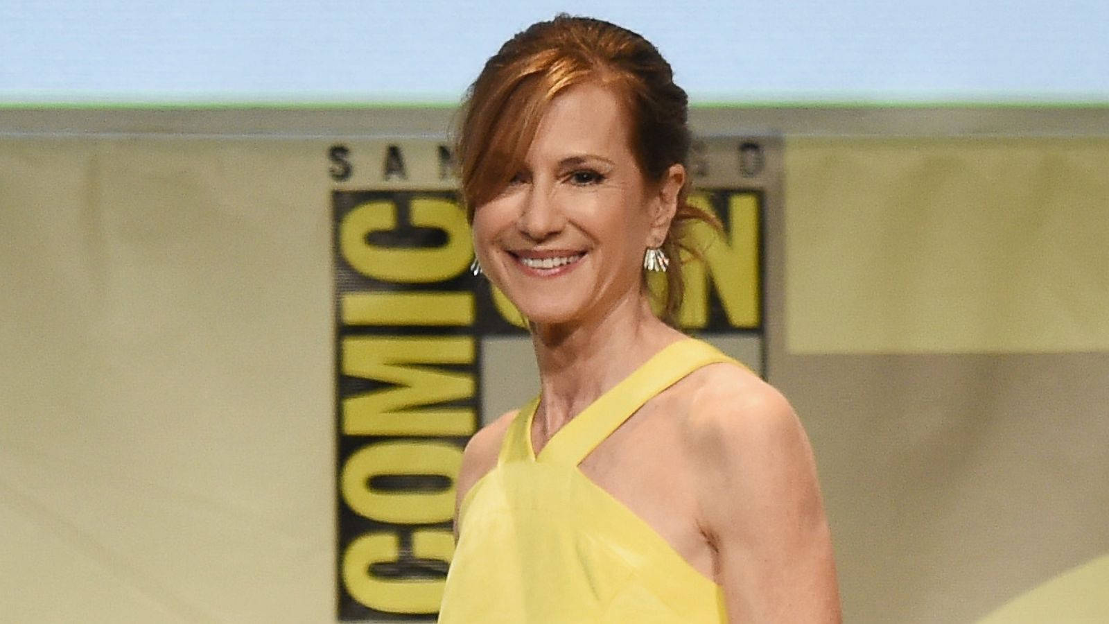 Celebrity 桌布 壁紙 — 一位身穿黃色連衣裙的微笑女士站在帶有粗體字樣的Comic-Con背景前。