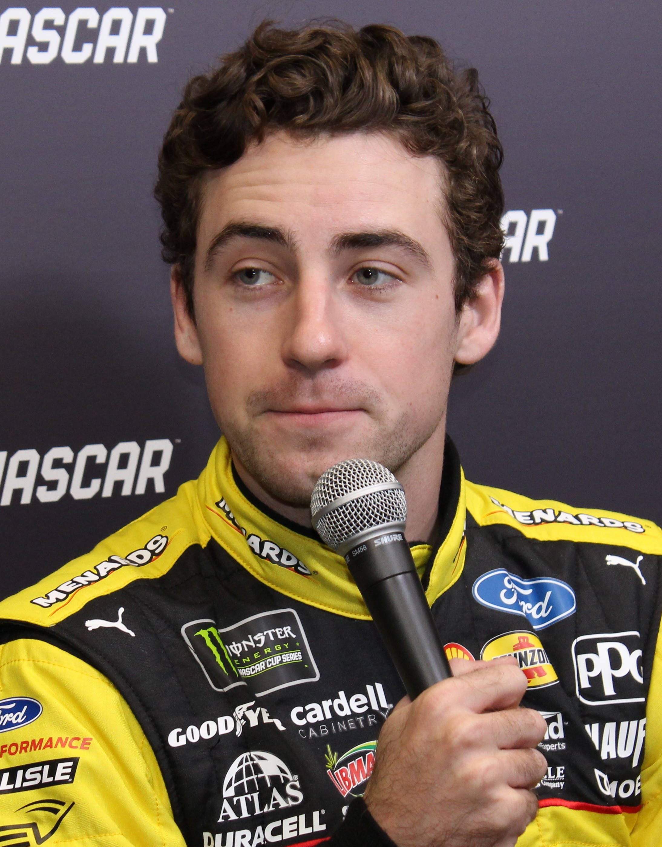 Ryan Blaney 桌布 壁紙 — 一位身穿黃黑賽車服的NASCAR車手在品牌背景前的新聞發布會上拿著麥克風。