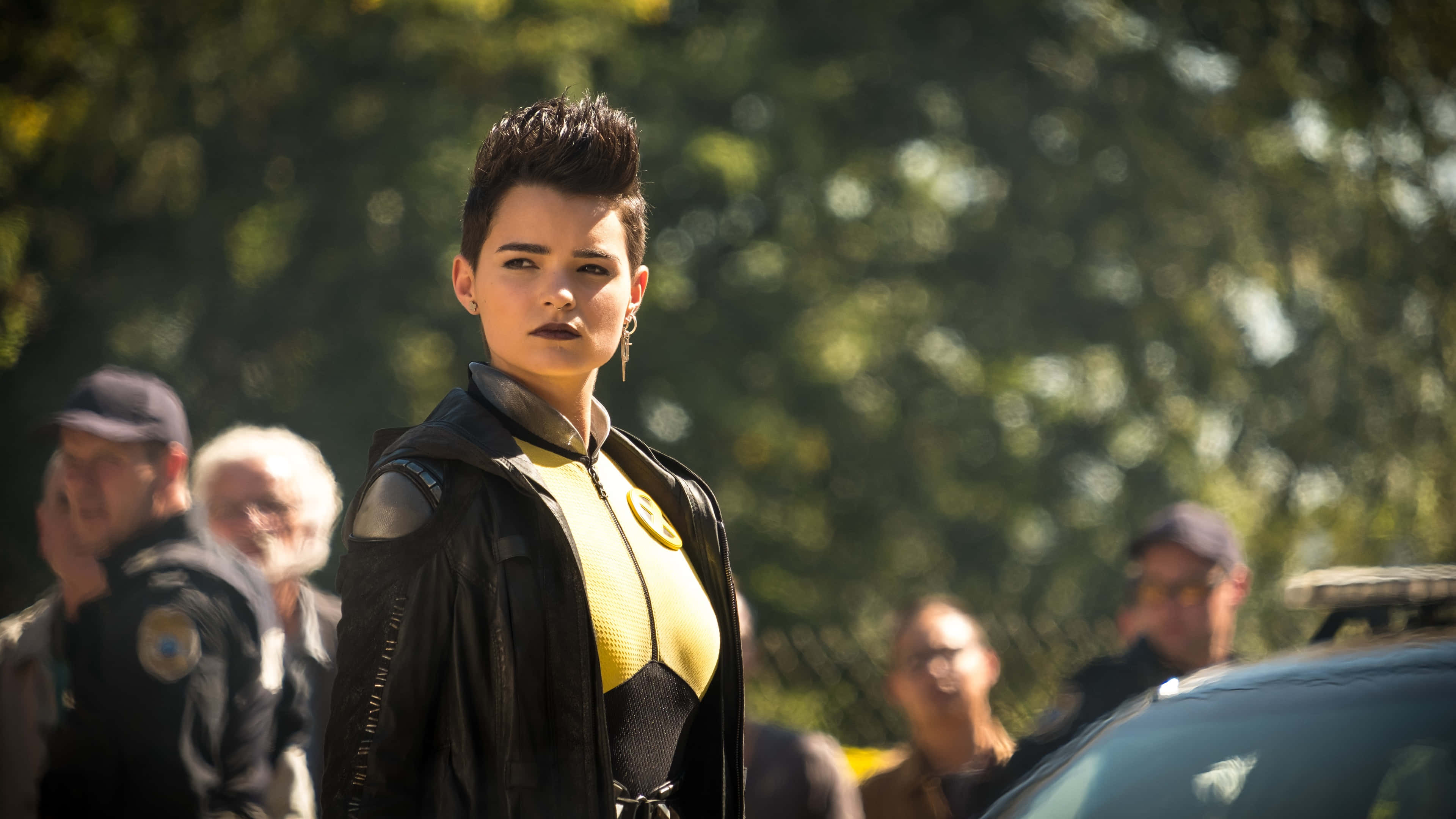 Negasonic Teenage Warhead 桌布 壁紙 — 一位身穿黃黑超級英雄服裝的兇猛女性自信地站在戶外，身後有模糊的警察和一輛汽車。