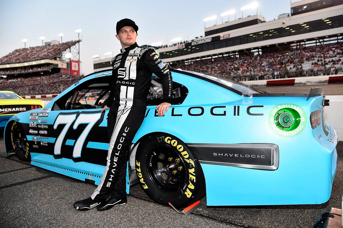 人物 桌布 壁紙 — 一位身穿黑白裝備的NASCAR車手在里奇蒙賽車場靠著他的淺藍色#77 ShaveLogic賽車，身後是擠滿觀眾的看台。