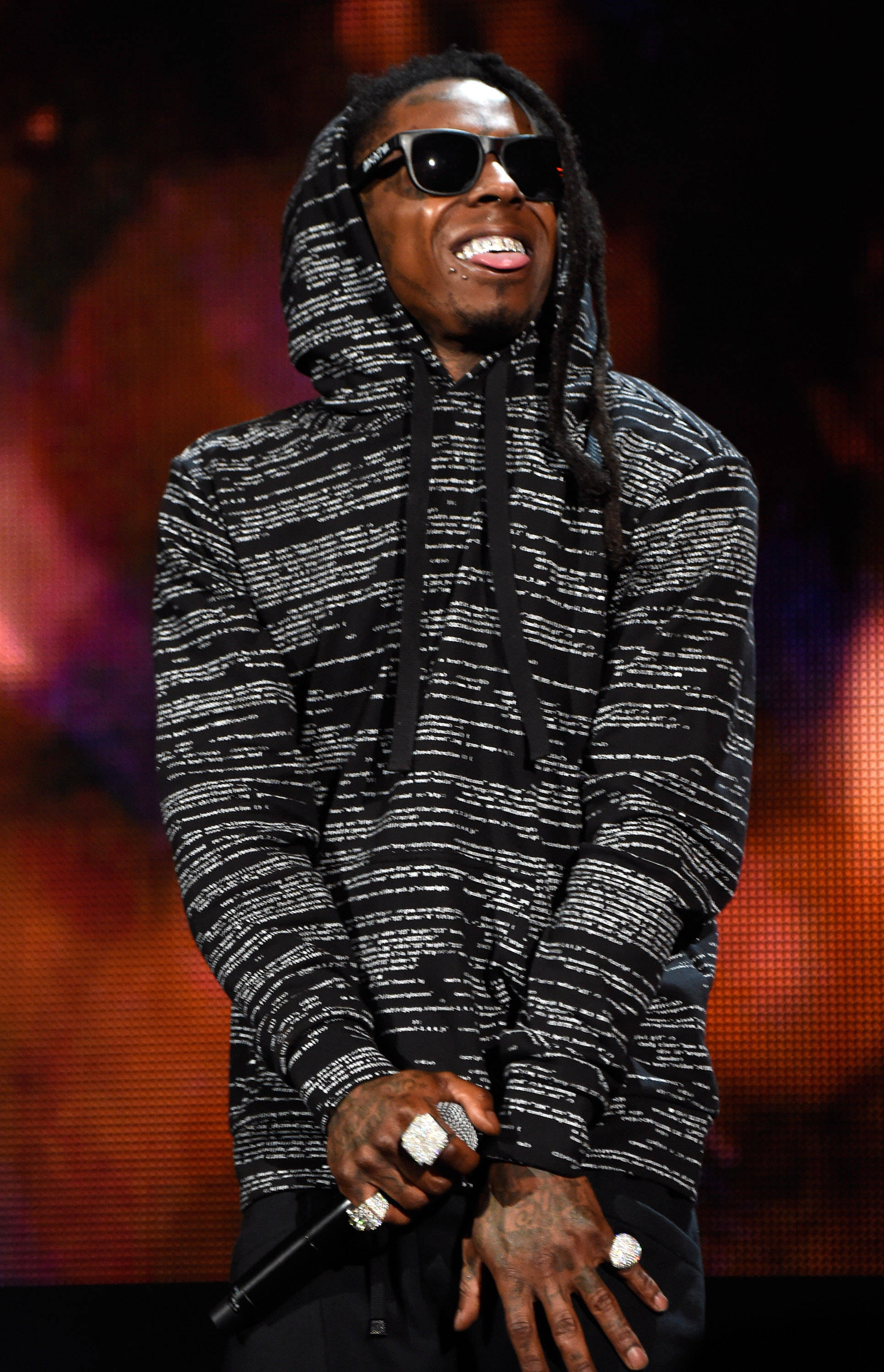 Lil Wayne 桌布 壁紙 — 一位身穿黑白連帽衫、戴著太陽眼鏡的饒舌歌手在燈光閃耀的舞台上咧嘴笑著，手握麥克風。