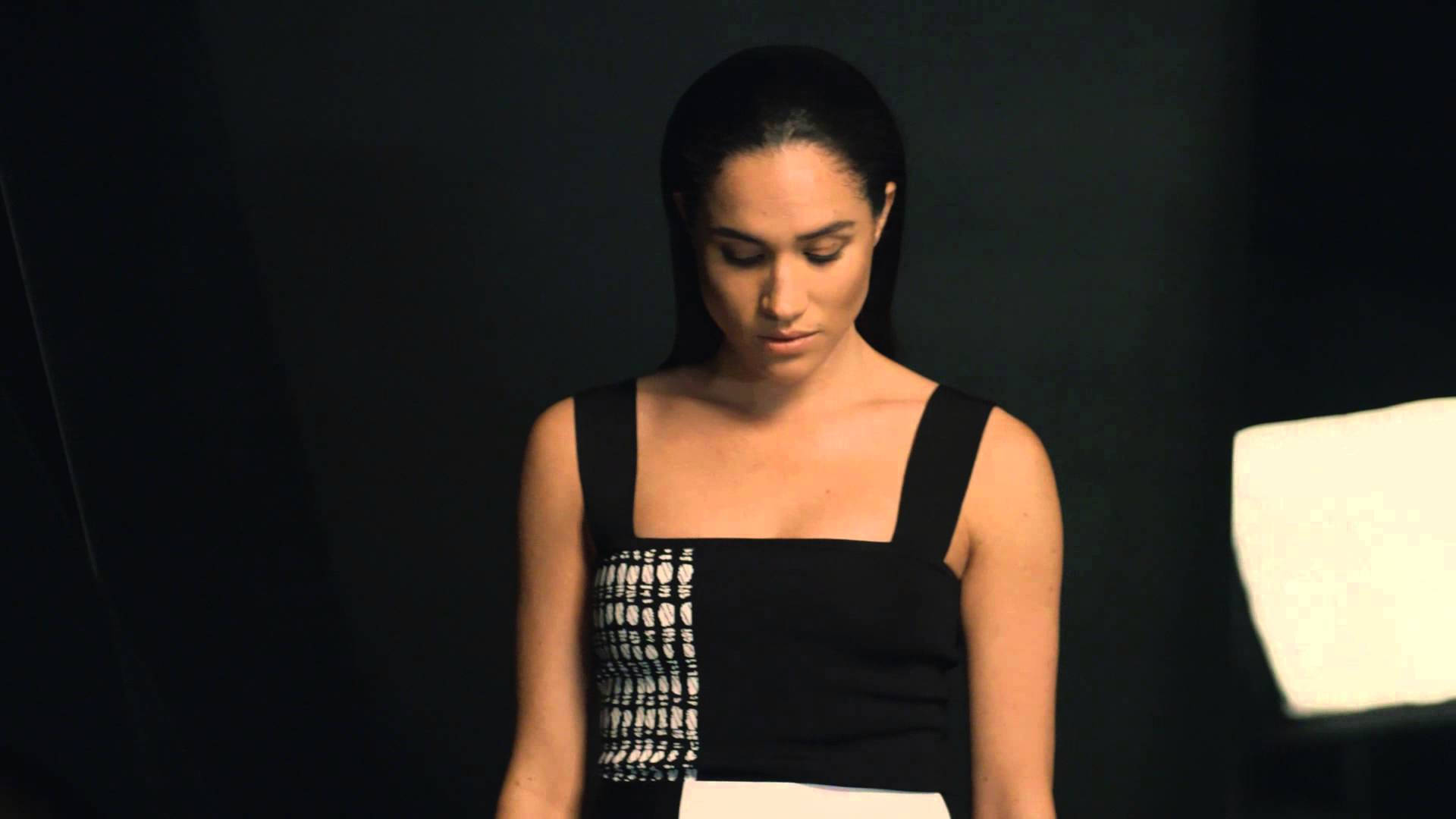Meghan Markle 桌布 壁紙 — 一位身穿黑白連衣裙的女性站在深色背景前，柔和的攝影棚燈光凸顯了她向下凝視的目光。