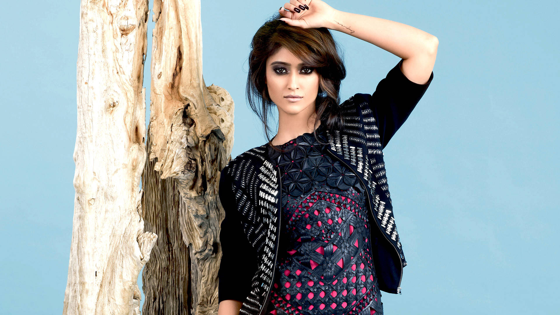 Ileana D'cruz 桌布 壁紙 — 一位身穿黑粉圖案服裝的女性，在純藍色背景前，自信地站在一棵風化的樹幹旁。