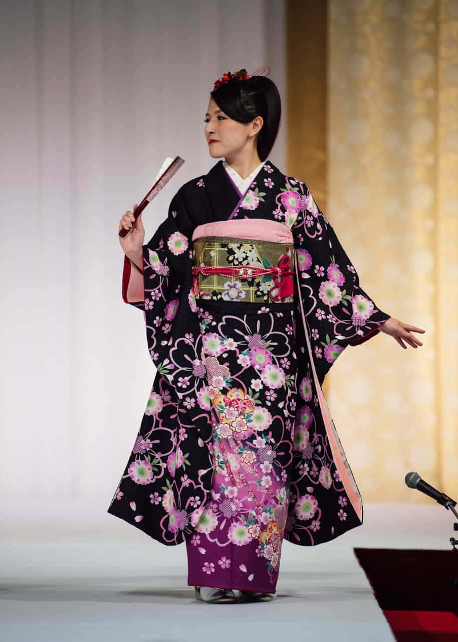 Kimono 桌布 壁紙 — 一位身穿黑粉花卉和服的女士優雅地在舞台上擺姿勢，手持扇子。