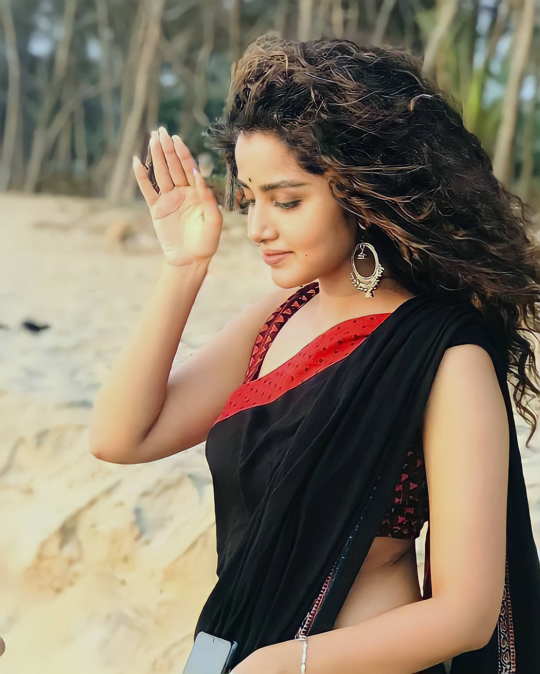 anupama parameswaran 桌布 壁紙 — 一位身穿黑紅色紗麗的女性站在沙灘上，以優雅的手勢遮擋陽光。