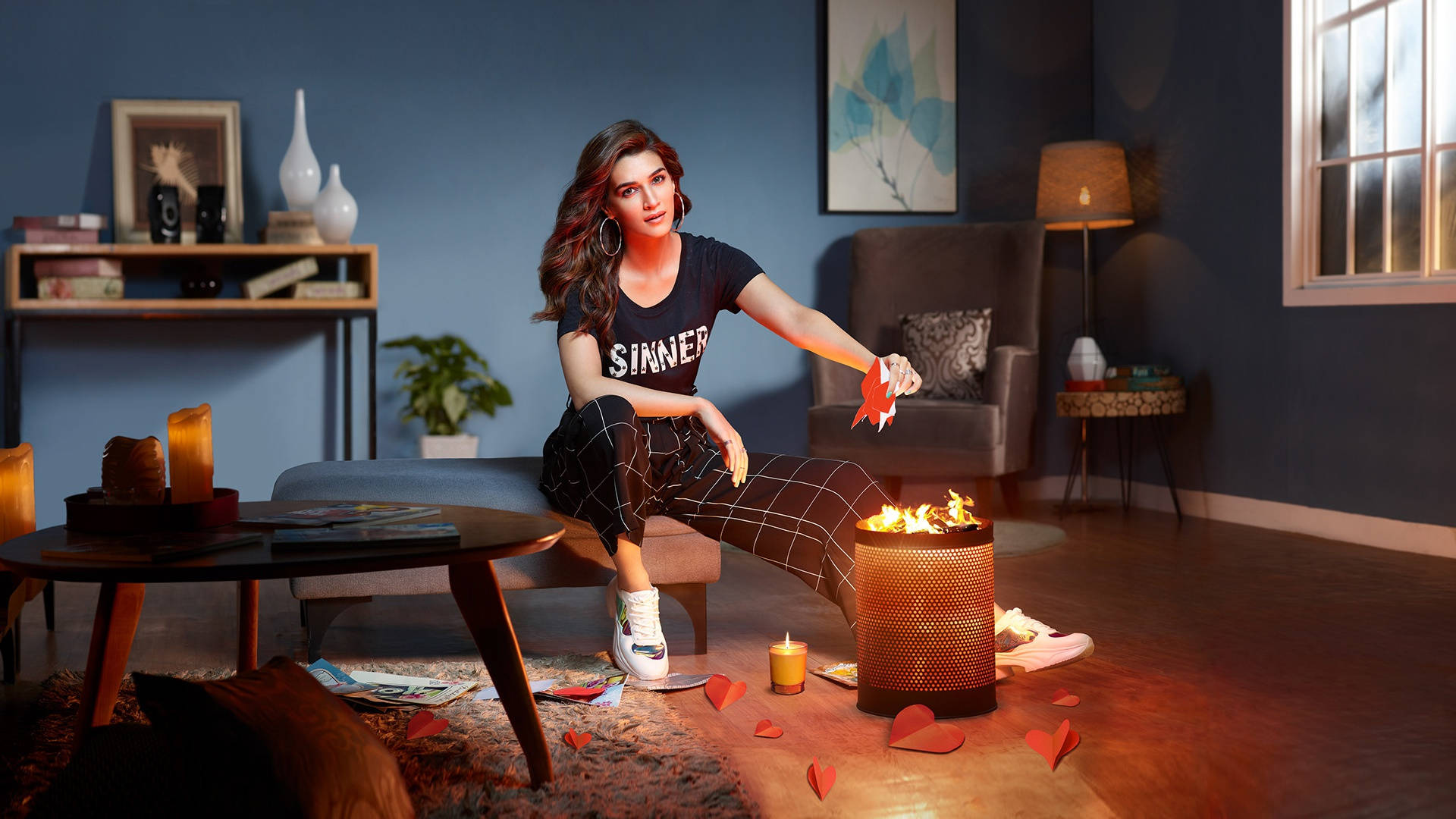 Kriti 桌布 壁紙 — 一位身穿黑色「SINNER」T恤的女性坐在昏暗藍色客廳的沙發上，在一個被散落的紅色紙心環繞的金屬燃燒器中點燃心形火焰。