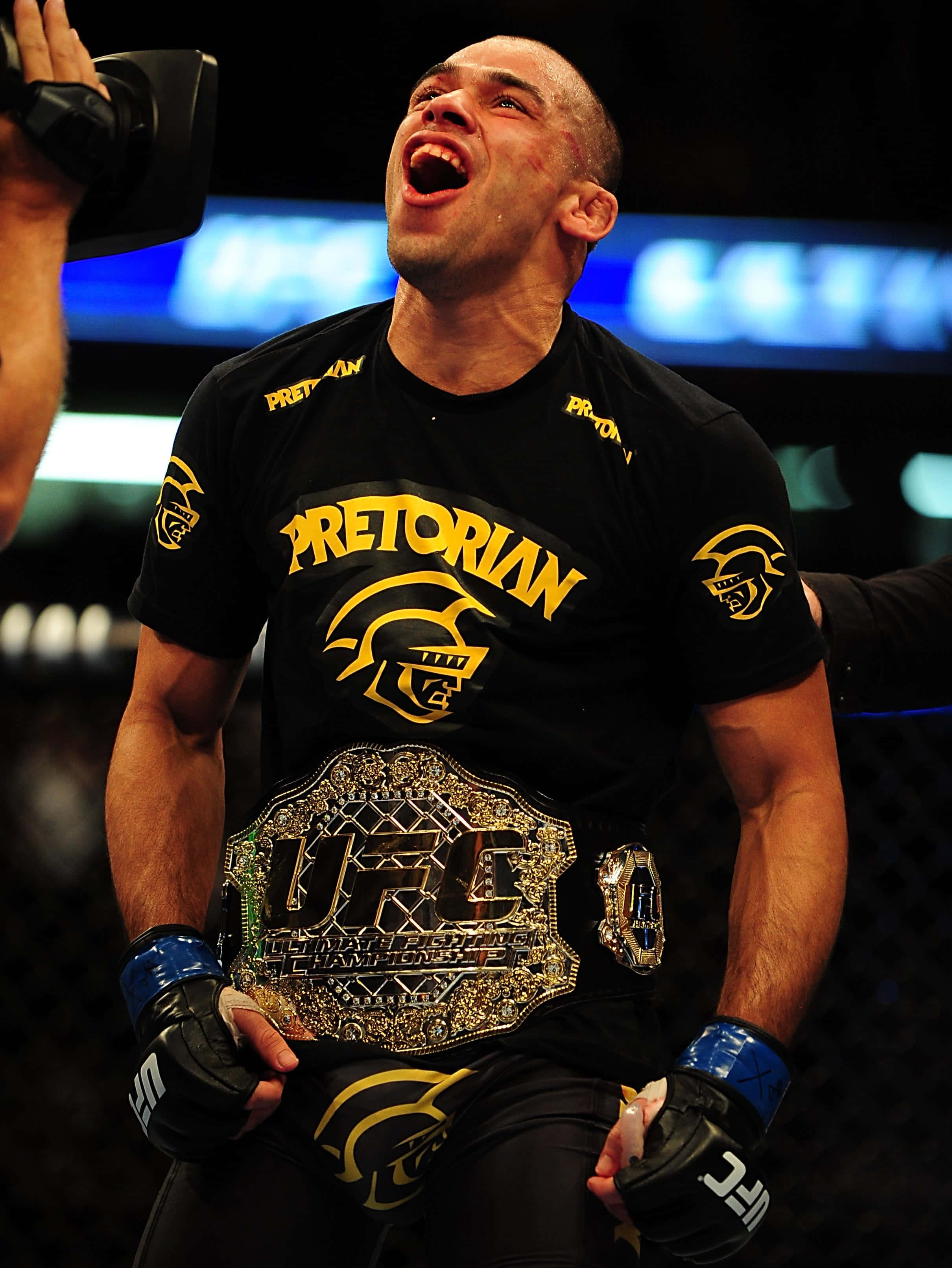 UFC 桌布 壁紙 — 一位身穿黑色Pretorian襯衫的UFC勝利戰士，高舉冠軍腰帶，發出勝利的怒吼。